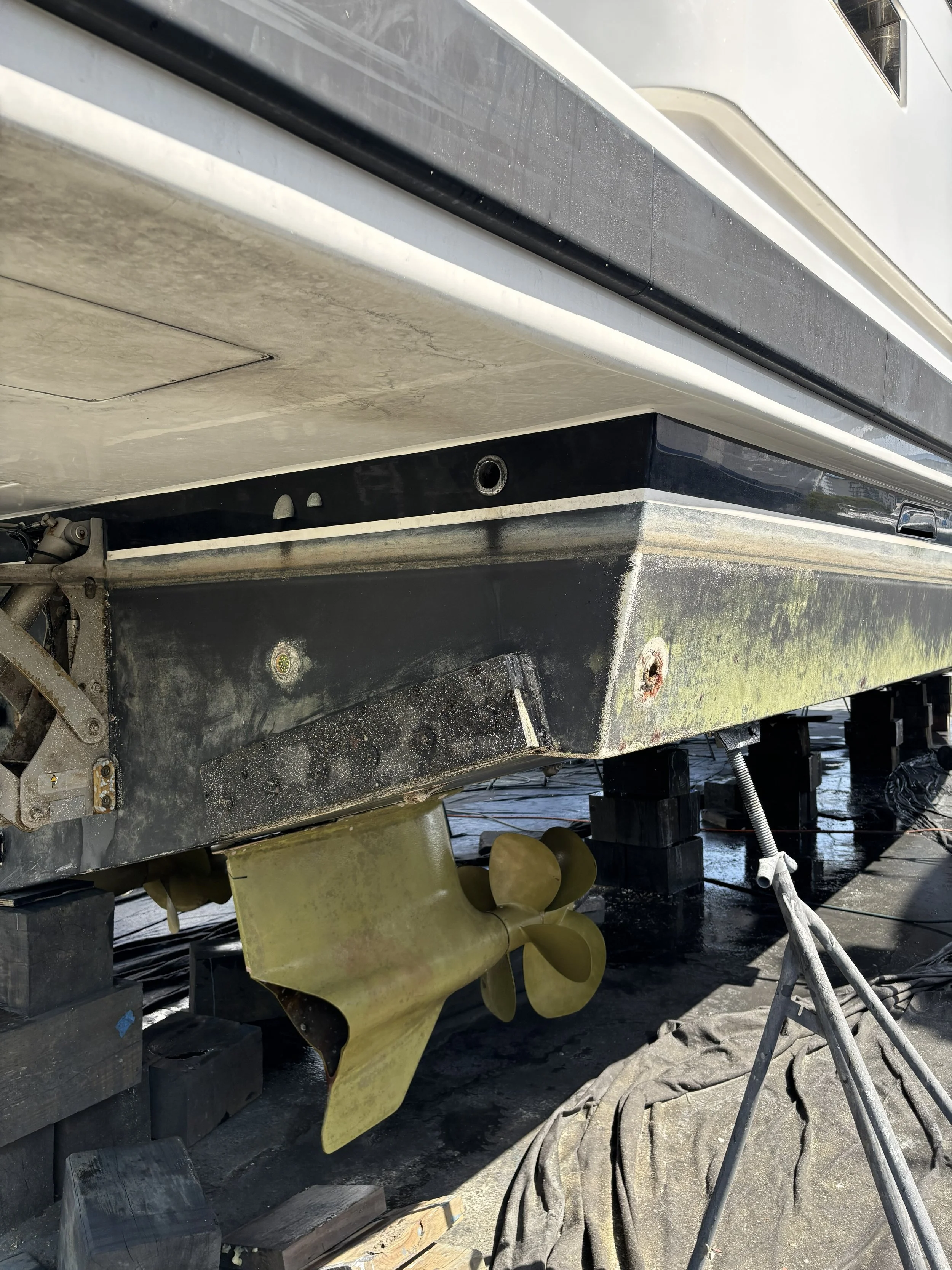 View of the underside of a boat on a trailer, with a yellow boat propeller visible, surrounded by black and yellow support blocks, and various ropes and equipment on the ground.