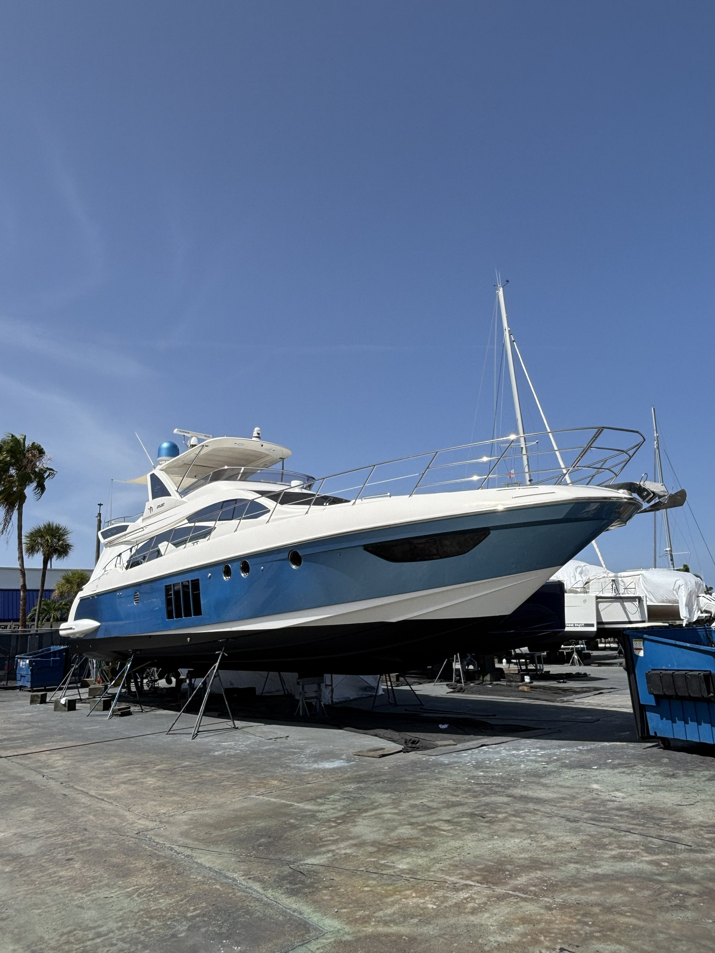 A large white and blue yacht is on a boat hoist in a marina yard, surrounded by other boats and marina equipment, with a clear blue sky overhead.