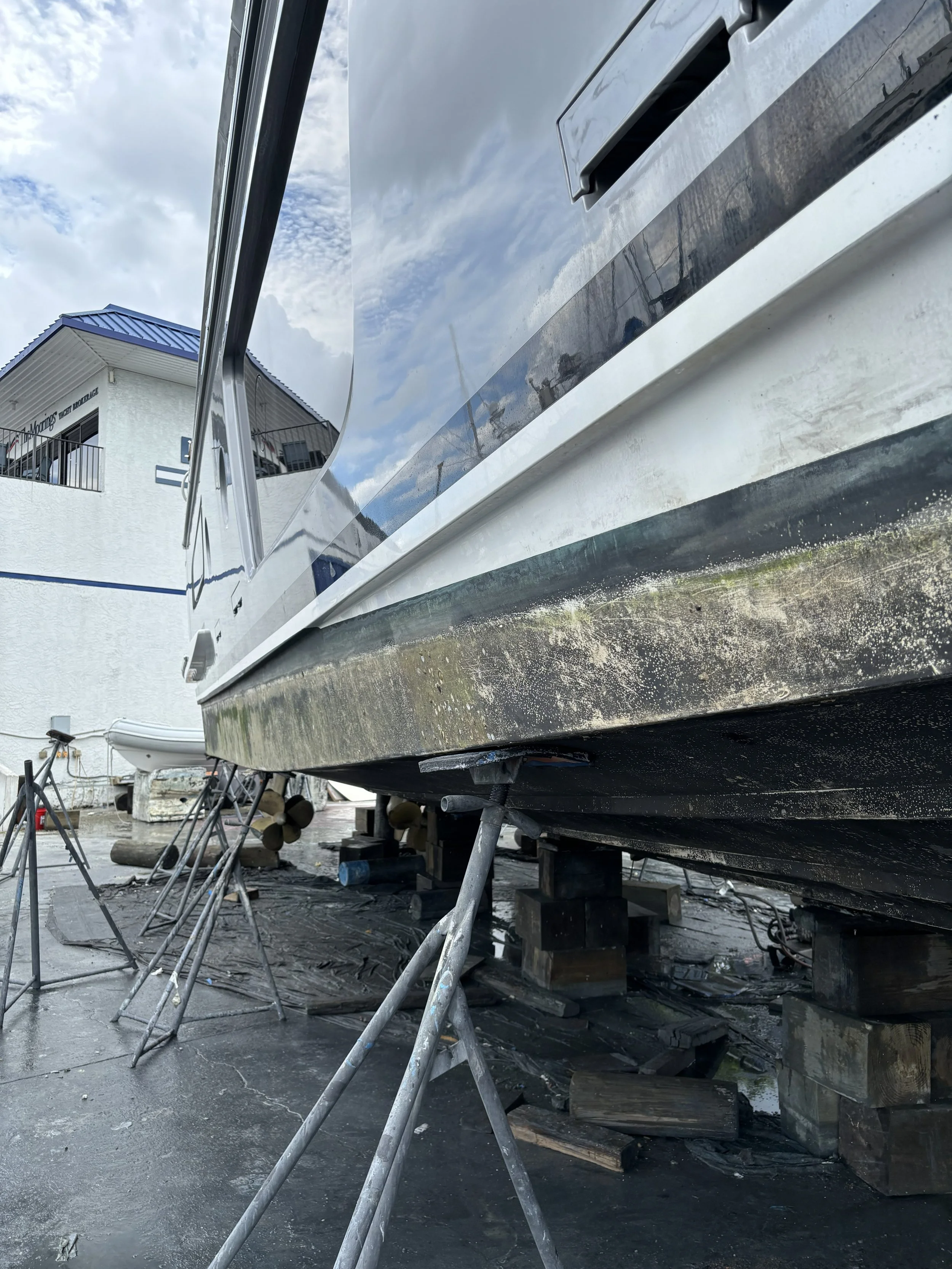 A boat is on a stand outside, with a building in the background. The boat's hull is black with a white stripe, and it appears to be under maintenance or storage.