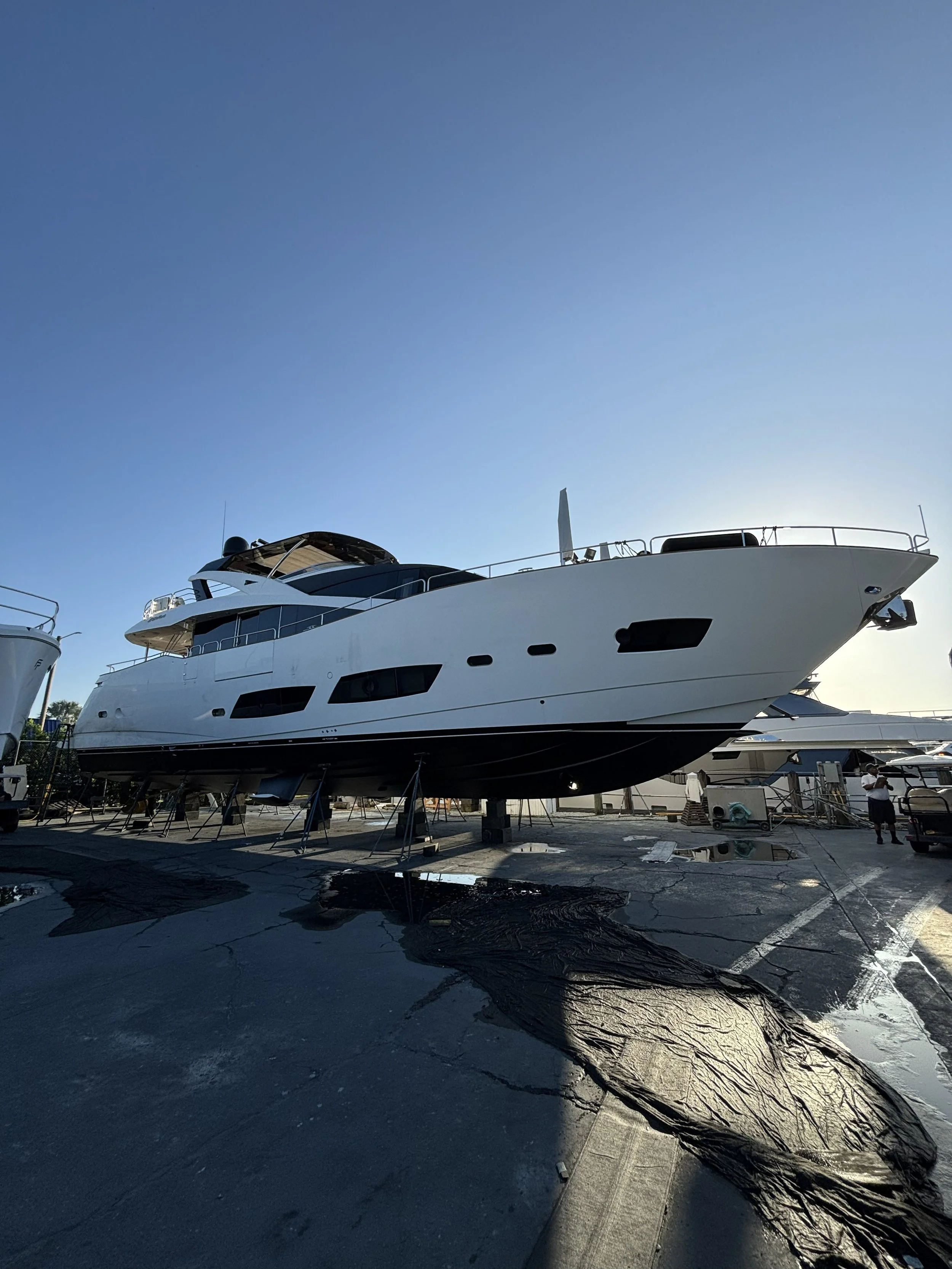 Large white yacht boat on dry dock for maintenance, with workers and tools around its base, under clear sky and bright sunlight.