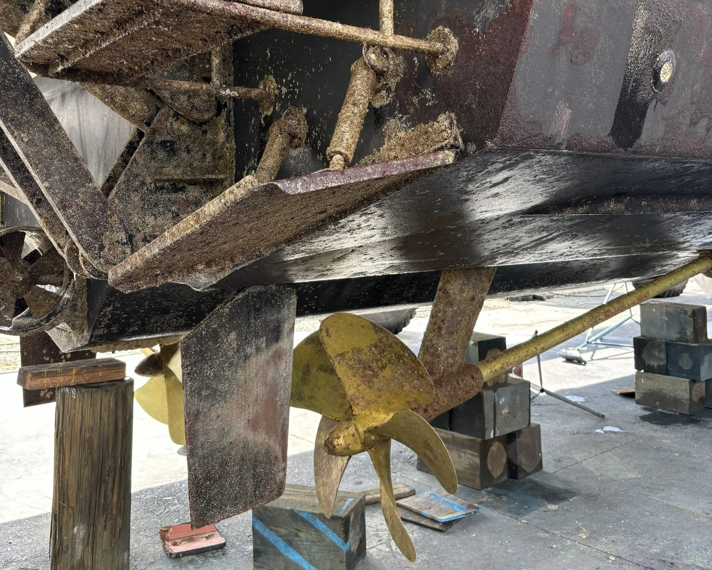 Close-up of a rusty, old piece of machinery or equipment, with various metal parts and joints, some covered in rust and grime, under a structure in an outdoor setting.