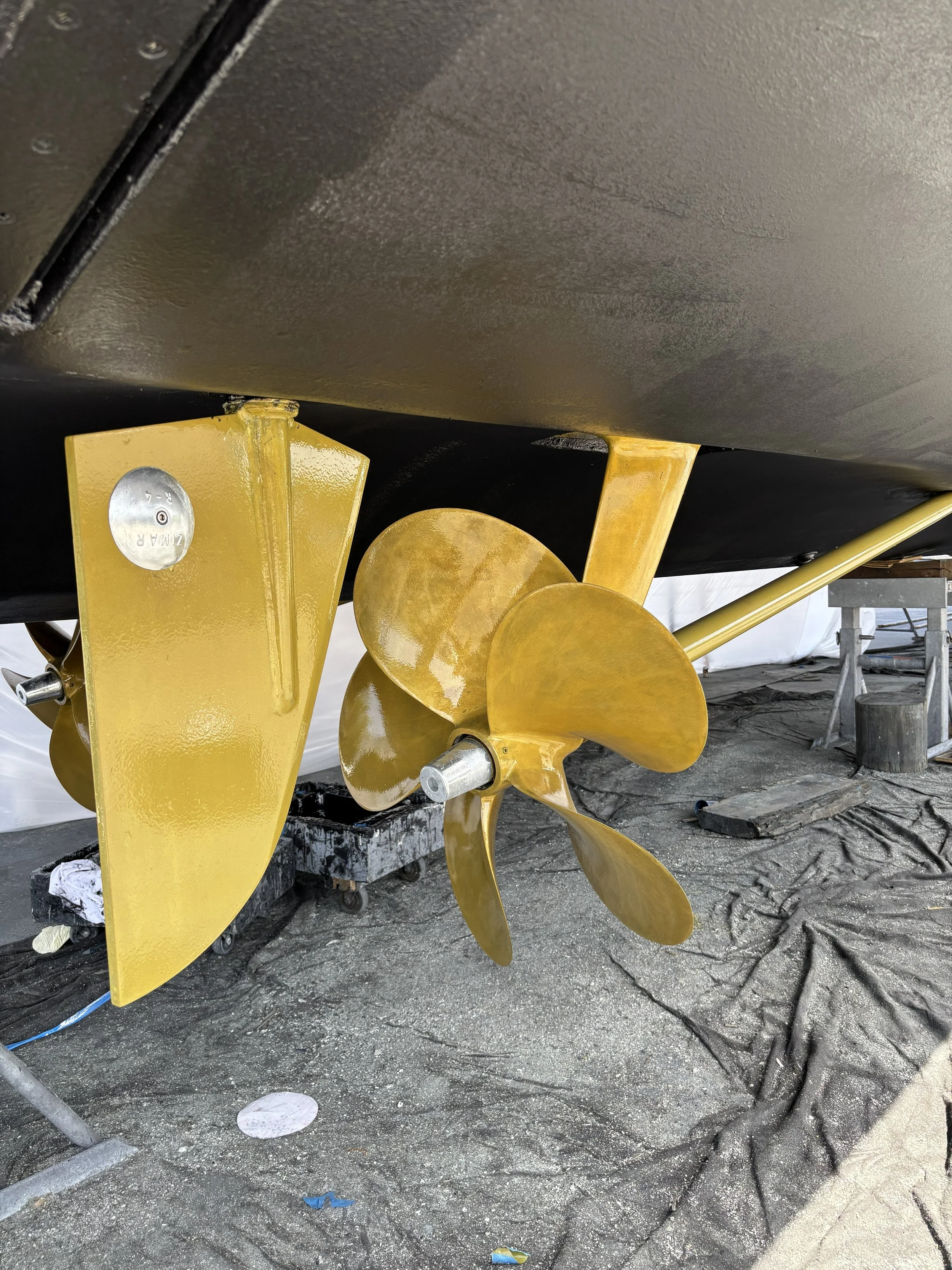 Underwater view of a boat's propeller and lower hull, showing the four-blade propeller and the boat's yellow-painted rudder and skeg. The boat is on a dry surface, possibly in a workshop or storage area.
