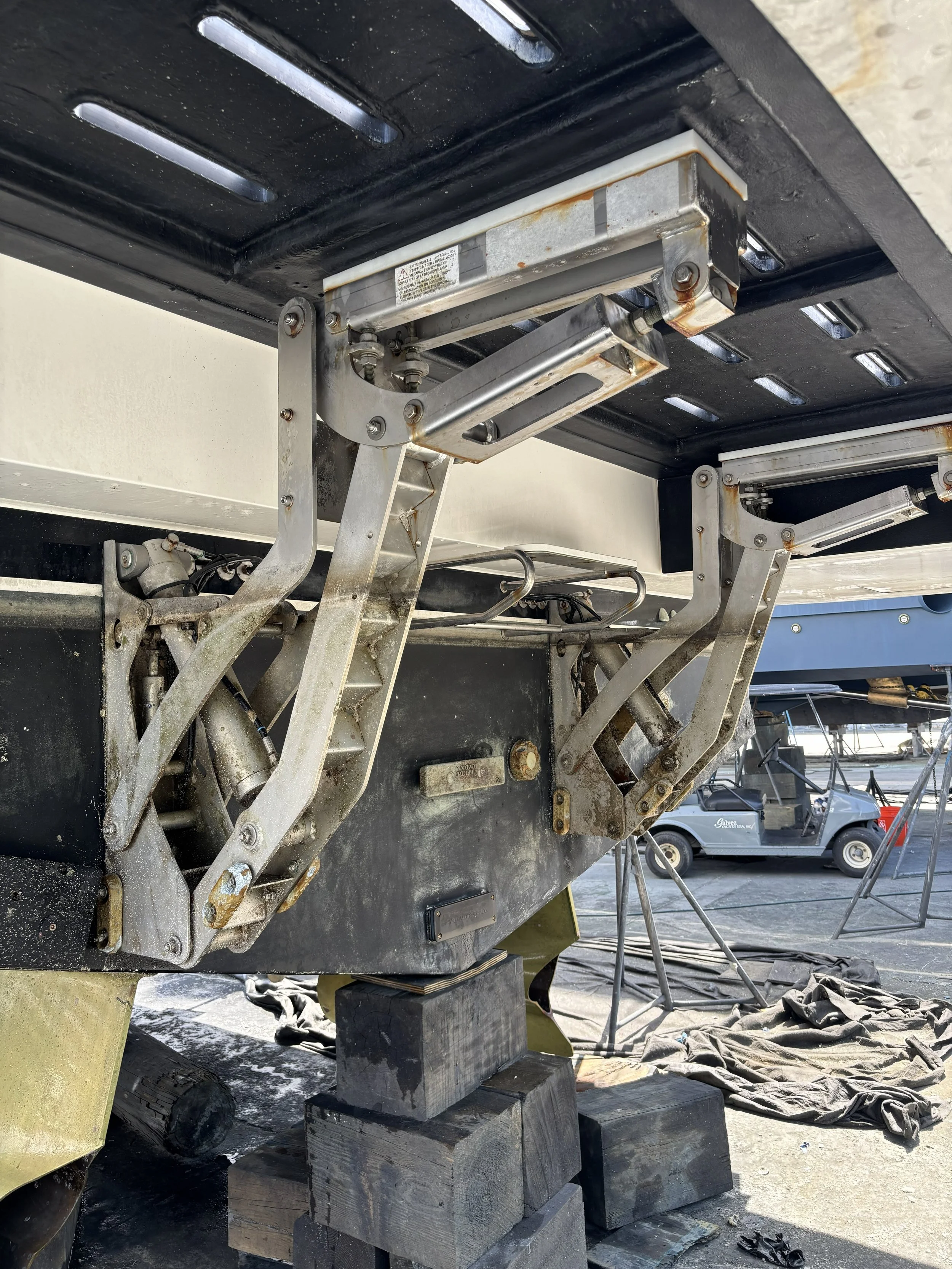 Close-up of a boat lift mechanism with metal arms and hydraulic parts, supporting a boat on wooden blocks during maintenance or construction in a boatyard.
