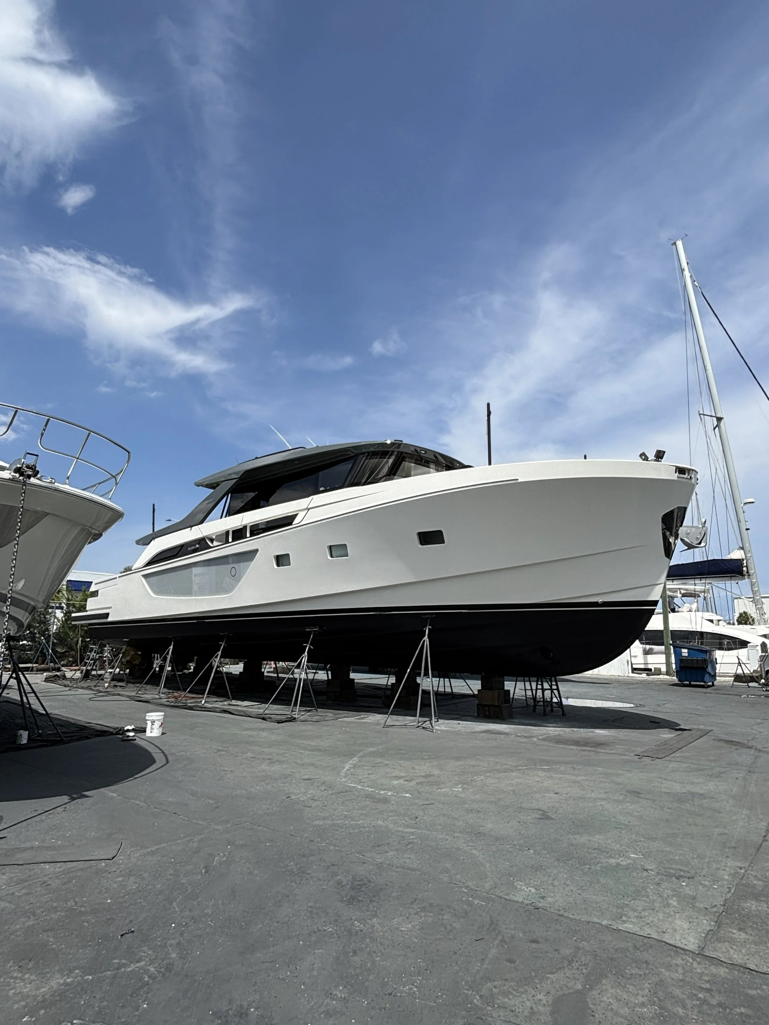 A large white yacht is on land, supported by wooden blocks and stands, with a blue sky and wispy clouds overhead.