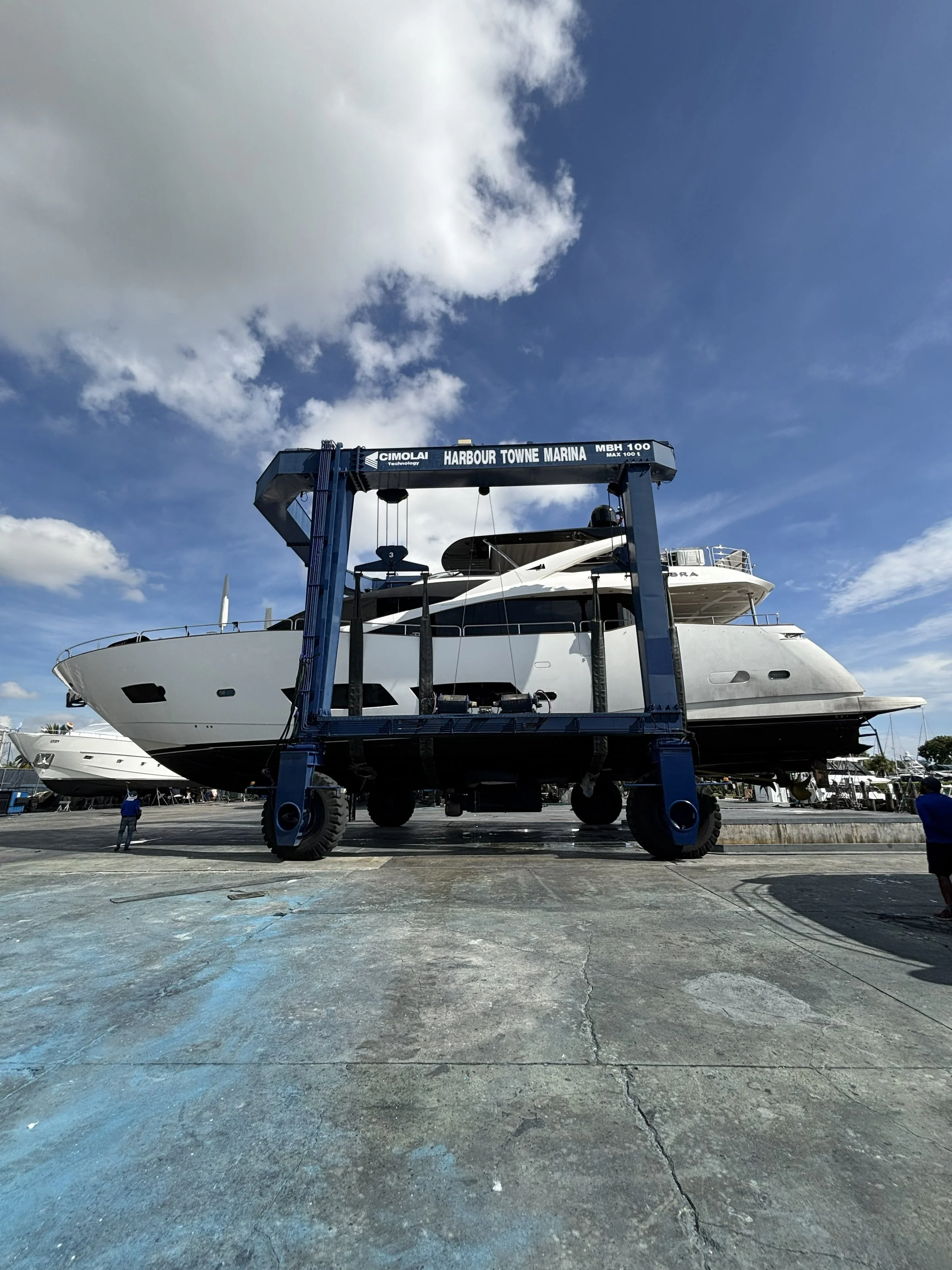 A large yacht is on a boat lift at a marina, with a blue sky and scattered clouds overhead.