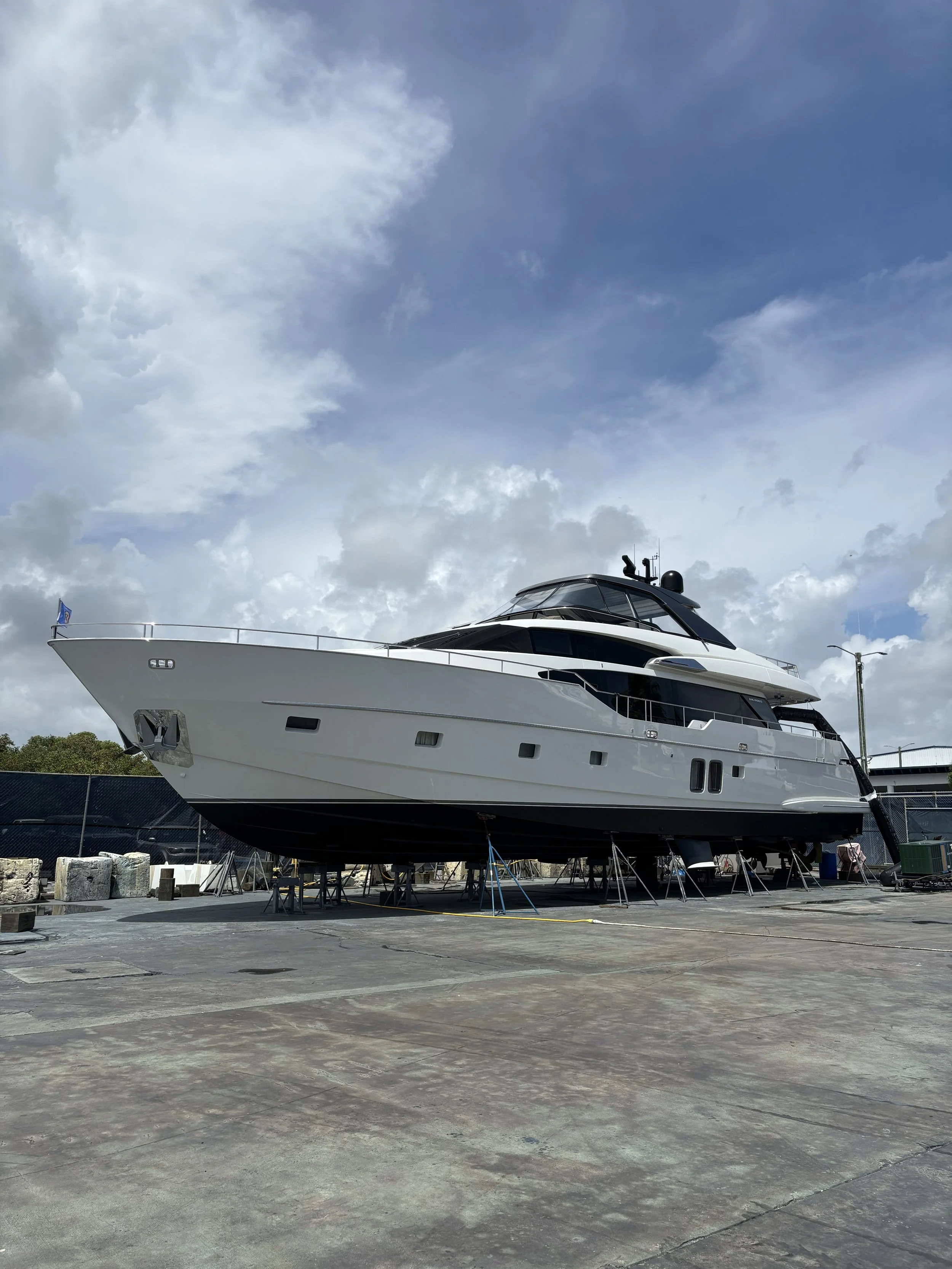 Large yacht on land in boatyard or shipyard, with support stands underneath, against cloudy sky.