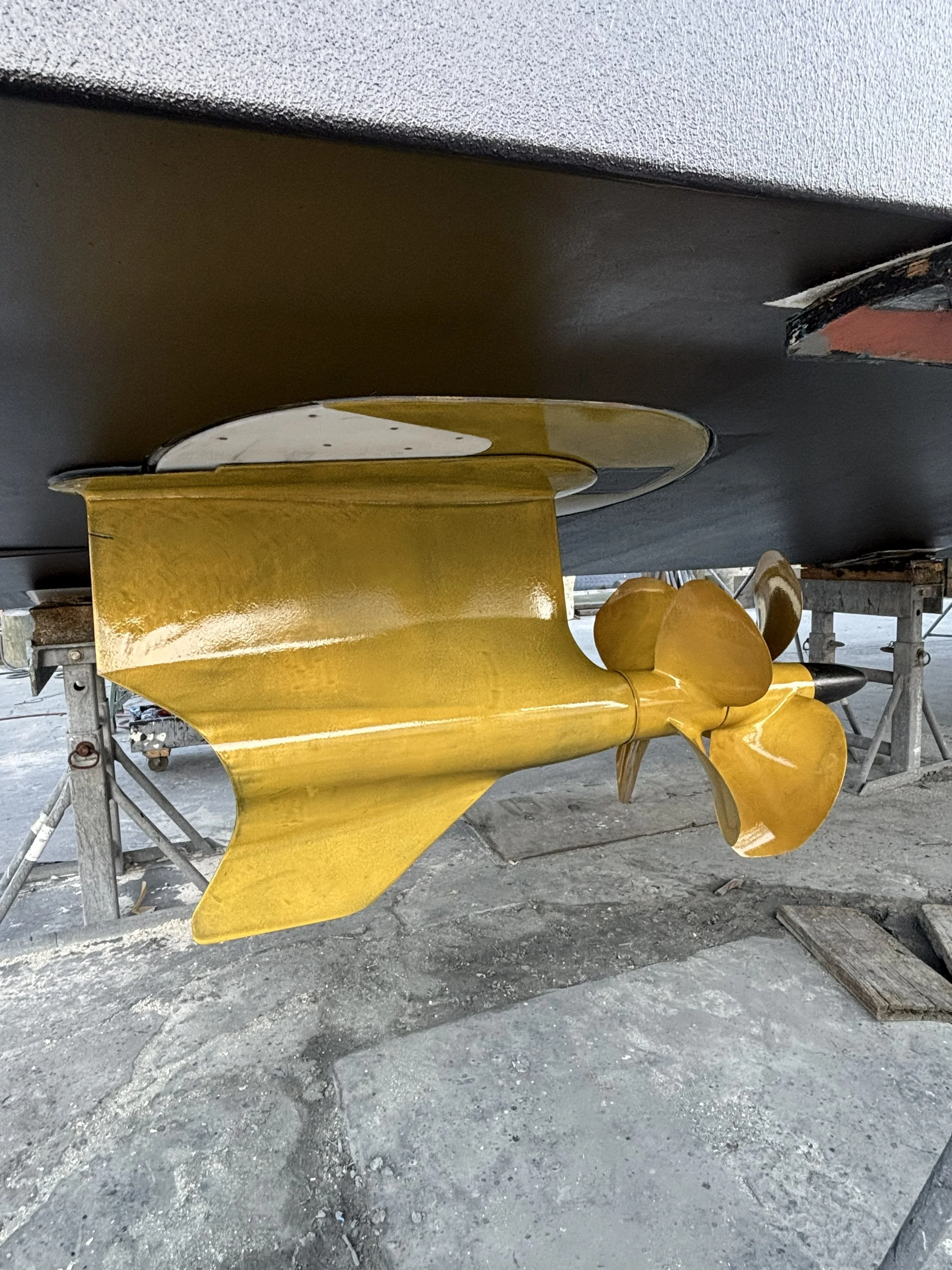 A boat with a yellow propeller and a yellow rudder, viewed from underneath, on a concrete surface.