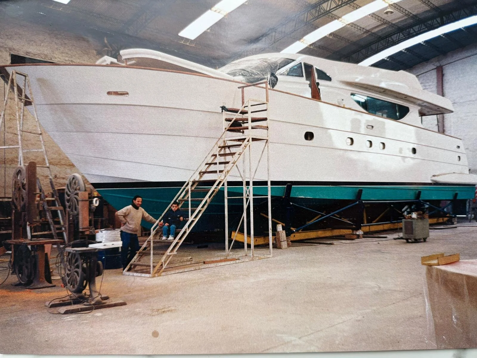 A large yacht under construction inside a manufacturing facility by engineers who are standing near the boat with equipment and scaffolding around them