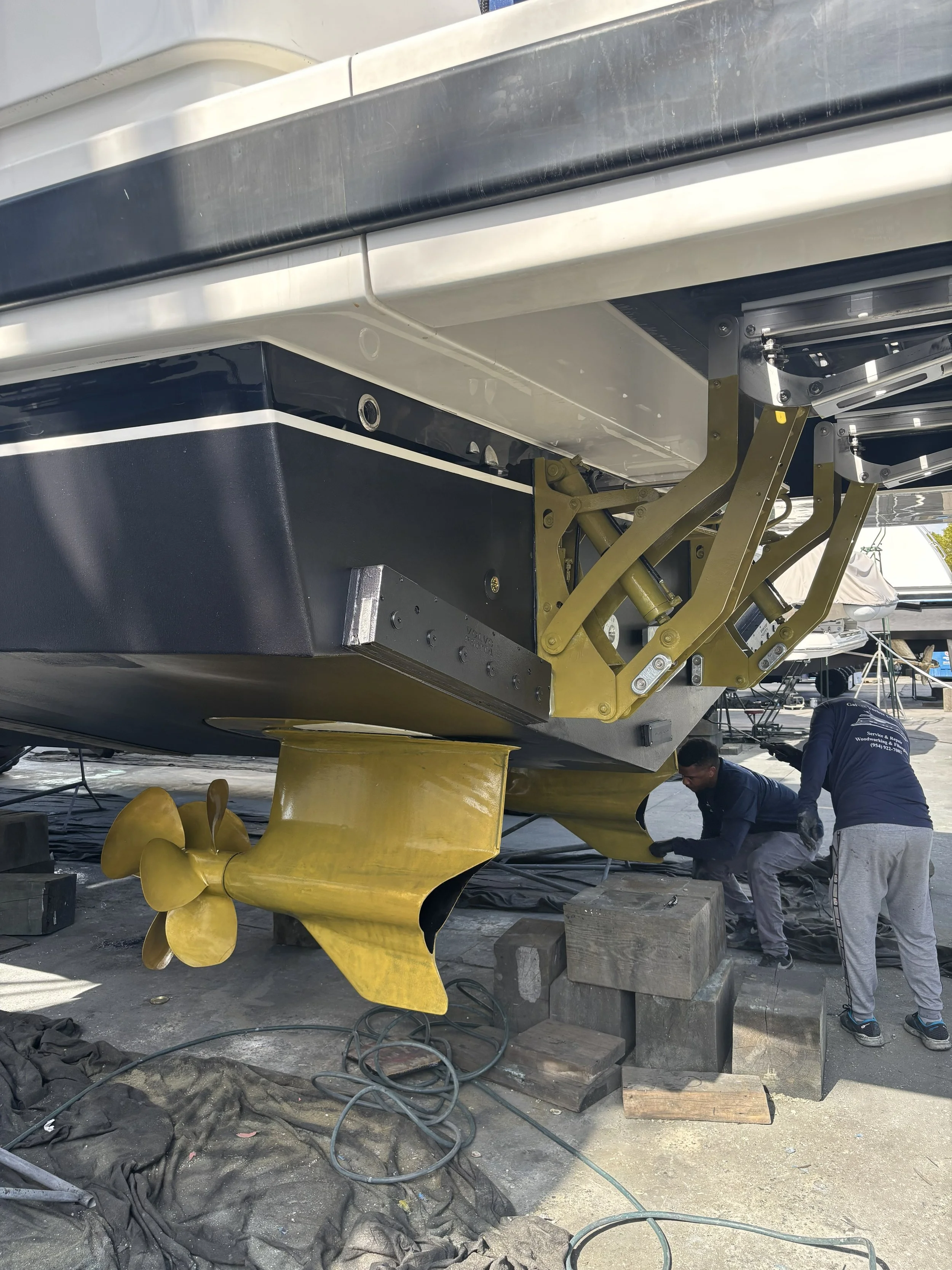 Two workers attach a large, yellow boat propeller to the underside of a boat while it is supported on blocks at a marina or shipyard.