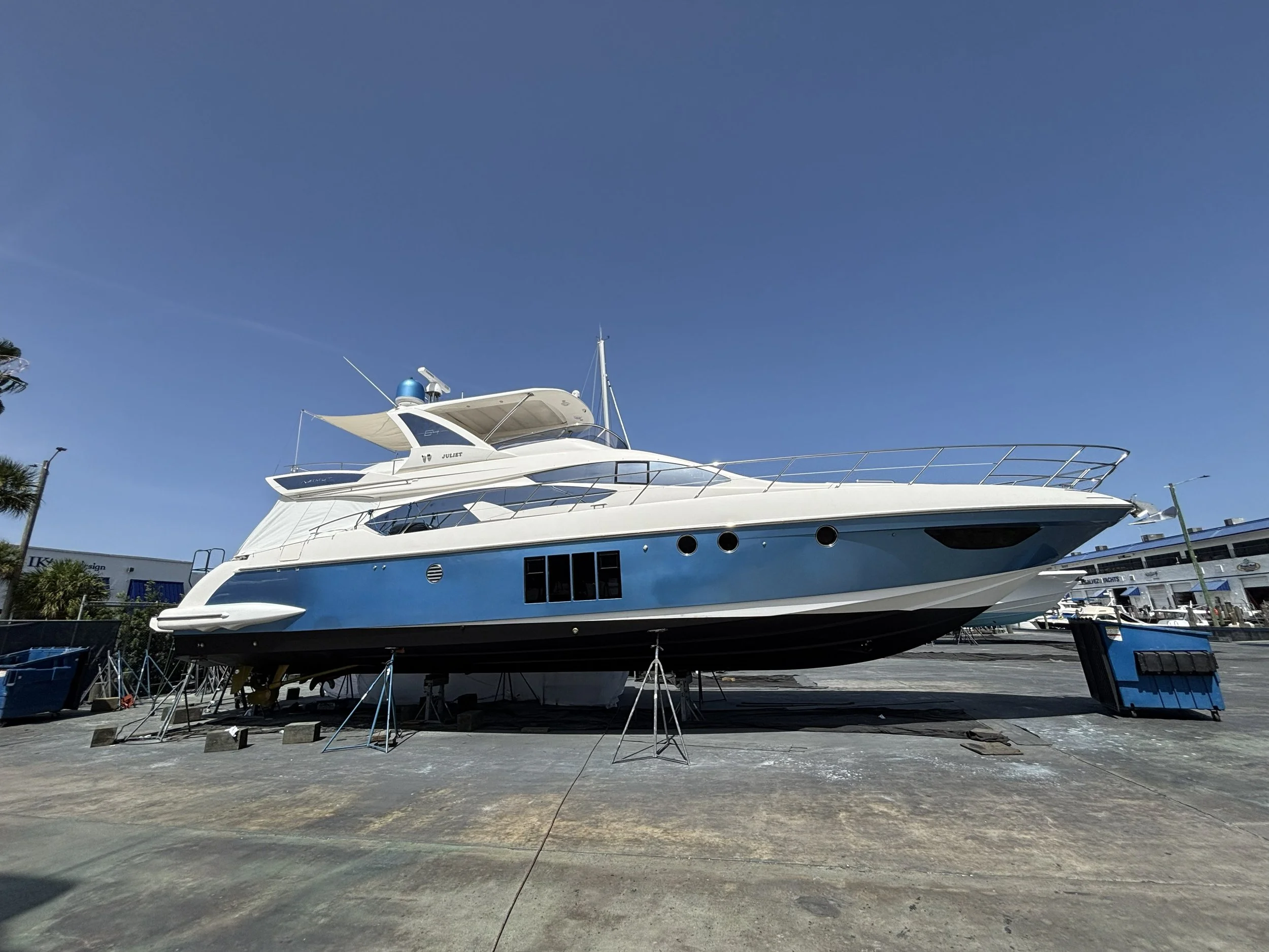 A white yacht with blue accents docked at a marina, with the name Juliet visible on the side. The yacht is under maintenance, supported by stands on a concrete lot, with a clear blue sky above.