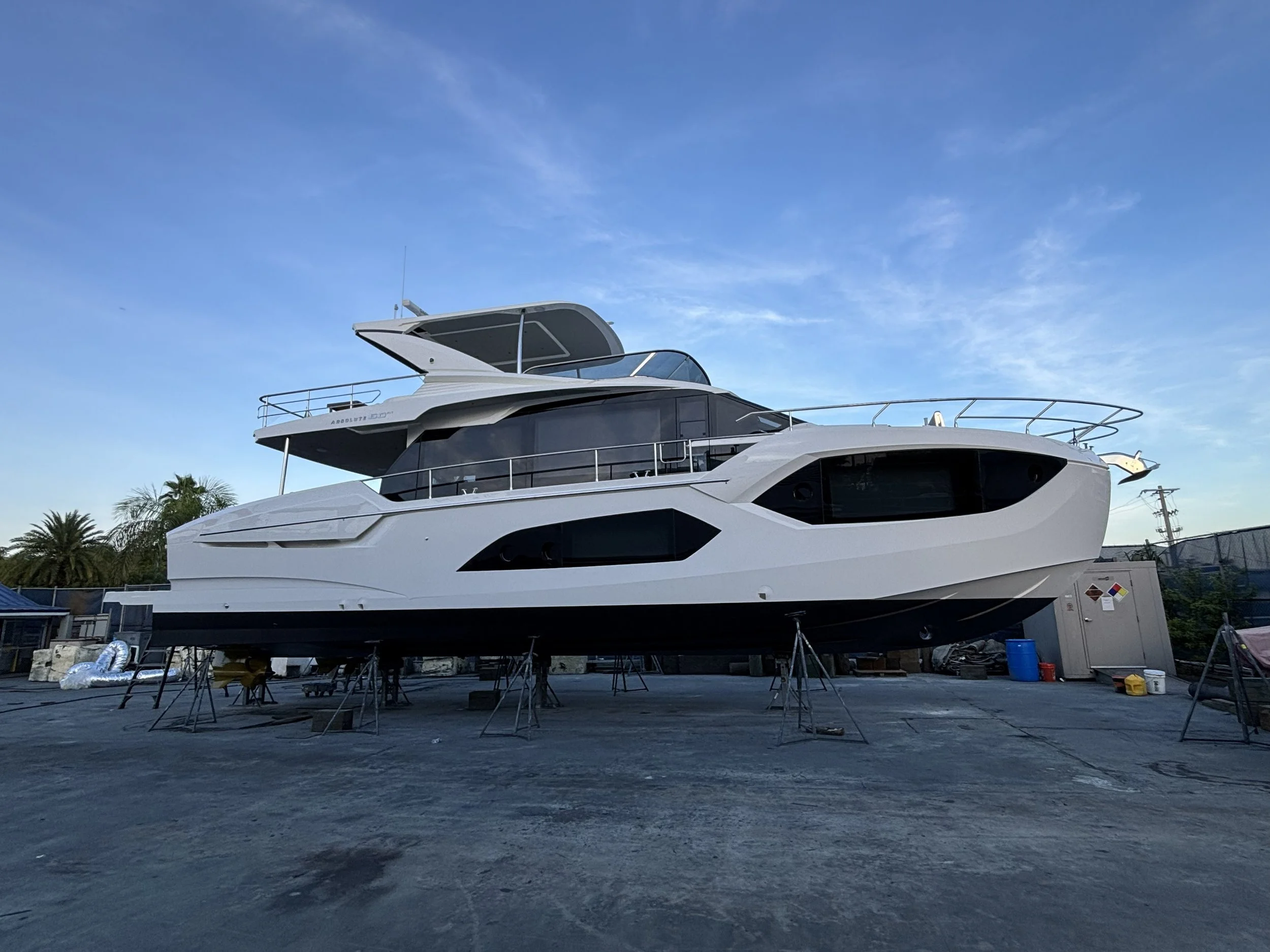 A large white yacht is on stands out of the water, in a boatyard. The boat has multiple decks, with the upper deck featuring a covered lounge area and railing. The sky is clear with a few wispy clouds, and there are some palm trees and power lines in
