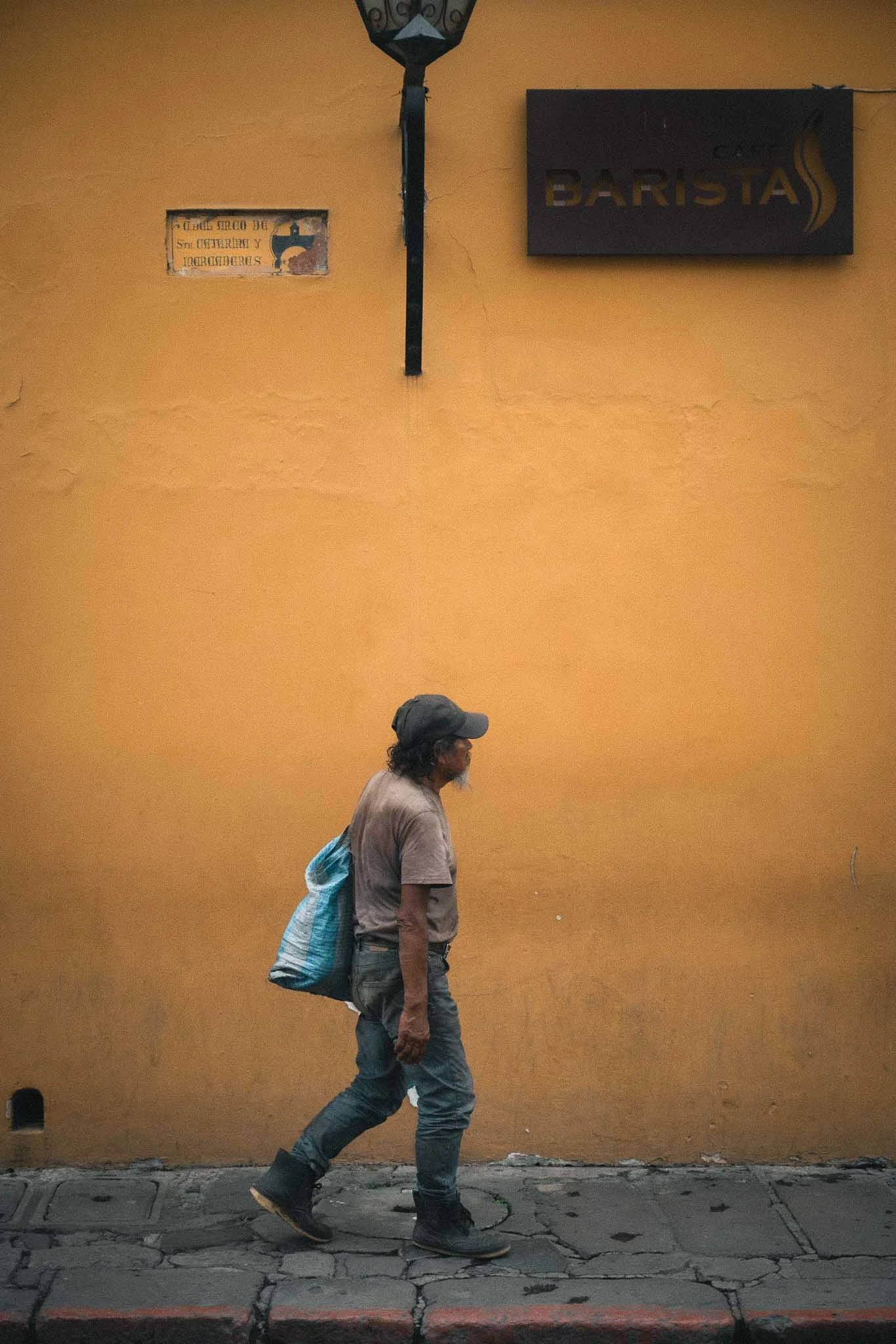 A man walking along a sidewalk in front of a yellow wall. The man is wearing a gray cap, a gray shirt, dark pants, and black boots. He is carrying a blue striped bag over his shoulder. There are two signs on the wall, one of which says "Barista".