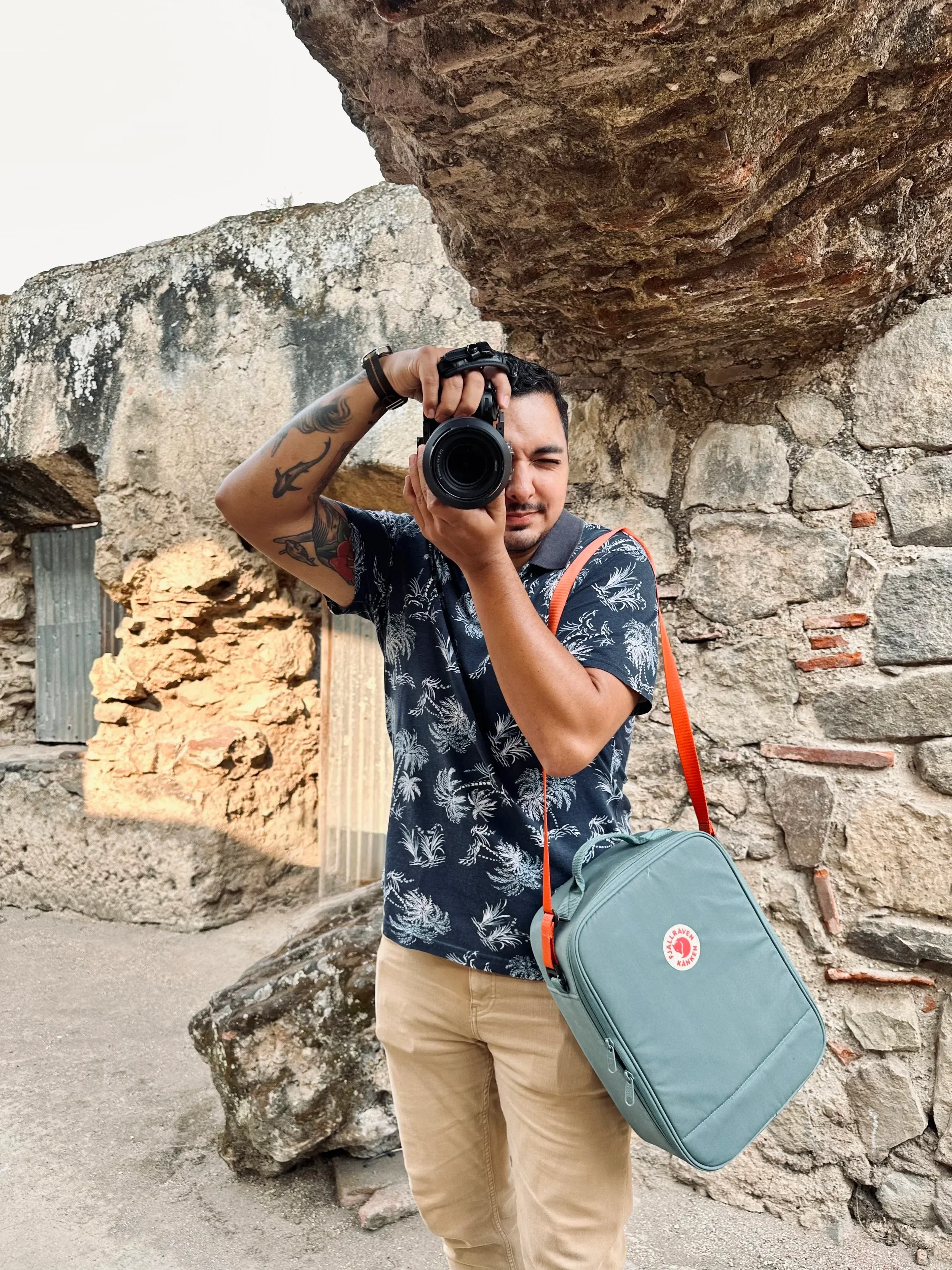 Man taking a photo with a camera in front of a weathered stone and brick structure outdoors. He is wearing a short-sleeve shirt, beige pants, and carrying a light blue bag with an orange strap.
