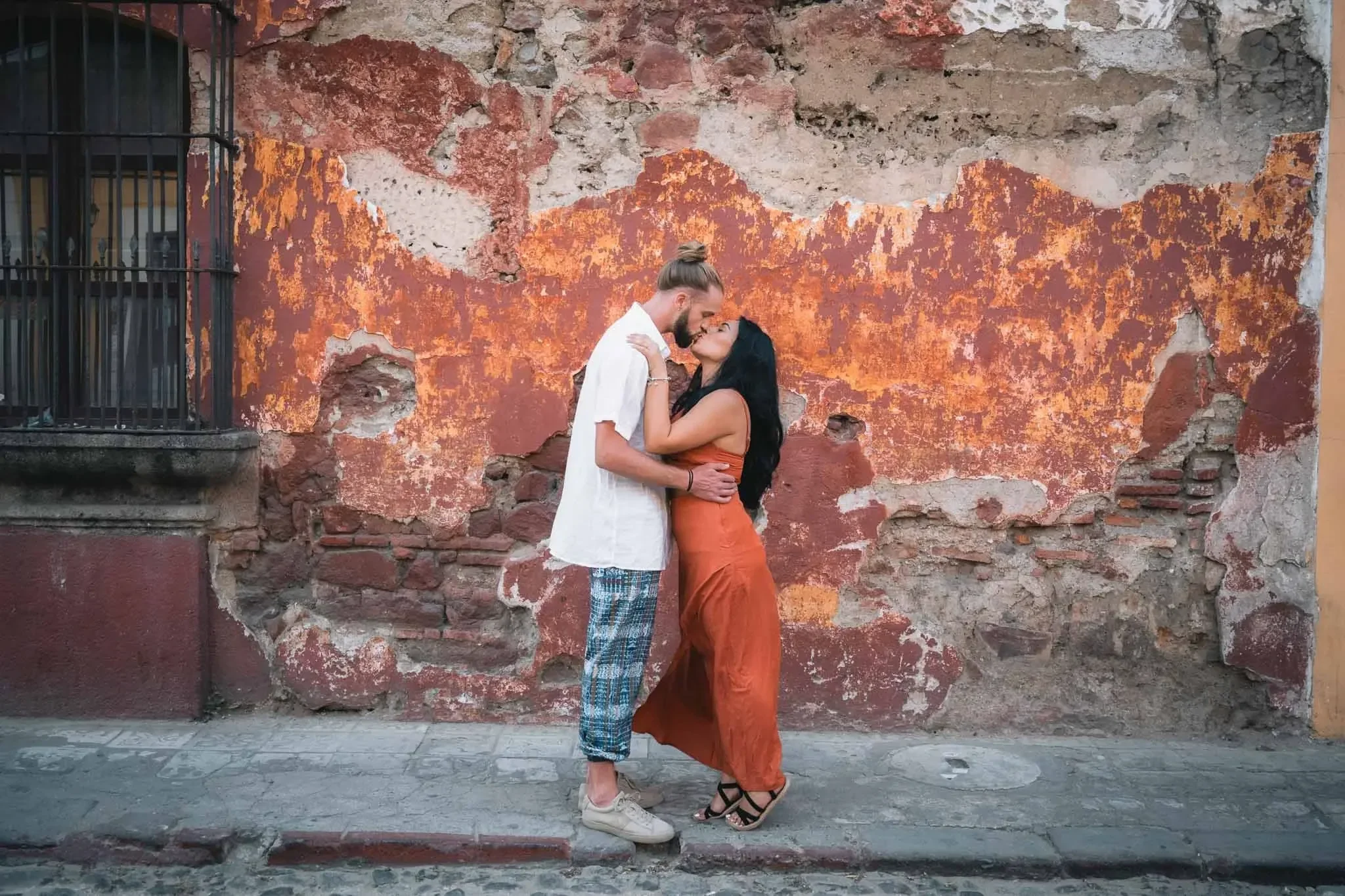 A couple is embracing and kissing in front of a weathered brick wall with peeling orange paint. The man has long hair tied back, a beard, and is wearing a white shirt and patterned pants. The woman has long black hair, wearing an orange dress and black high-heeled shoes.