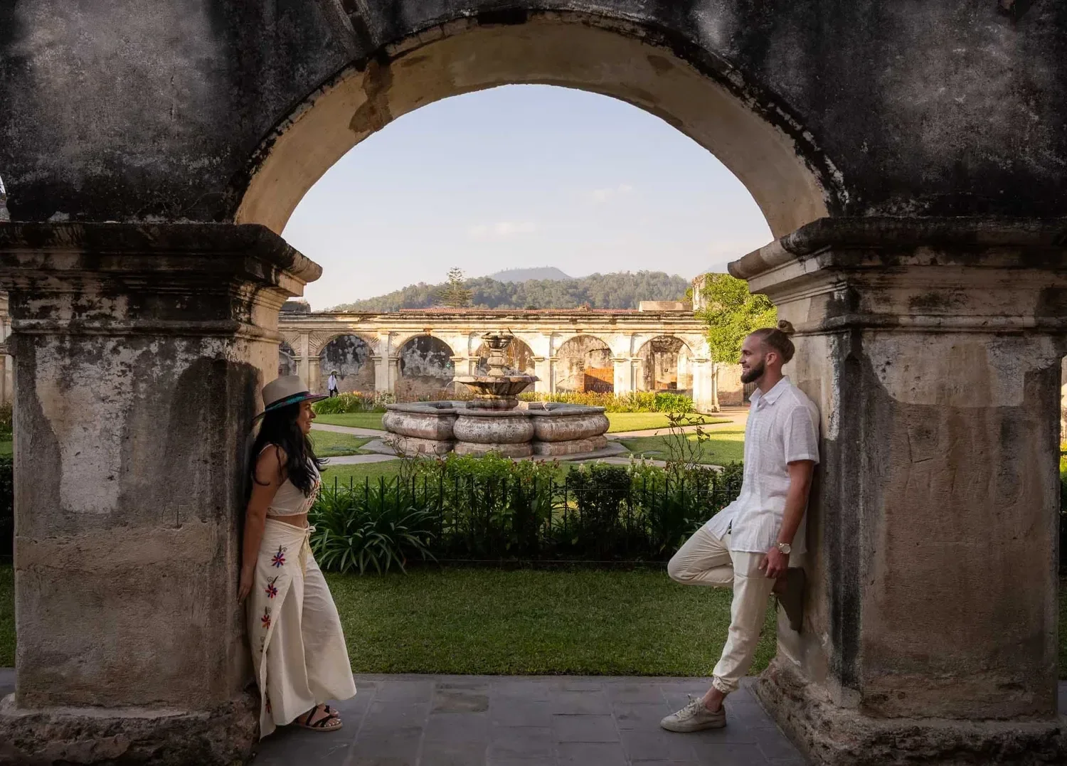 A man and woman standing under an old stone archway at a historic site or garden, facing each other and smiling, with a fountain and ancient ruins in the background.