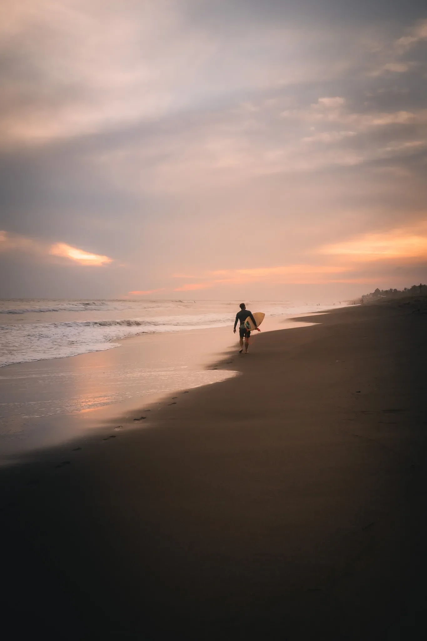 A person walking along the beach at sunset with a surfboard, the sky filled with soft clouds and the ocean waves gently crashing on the shore.