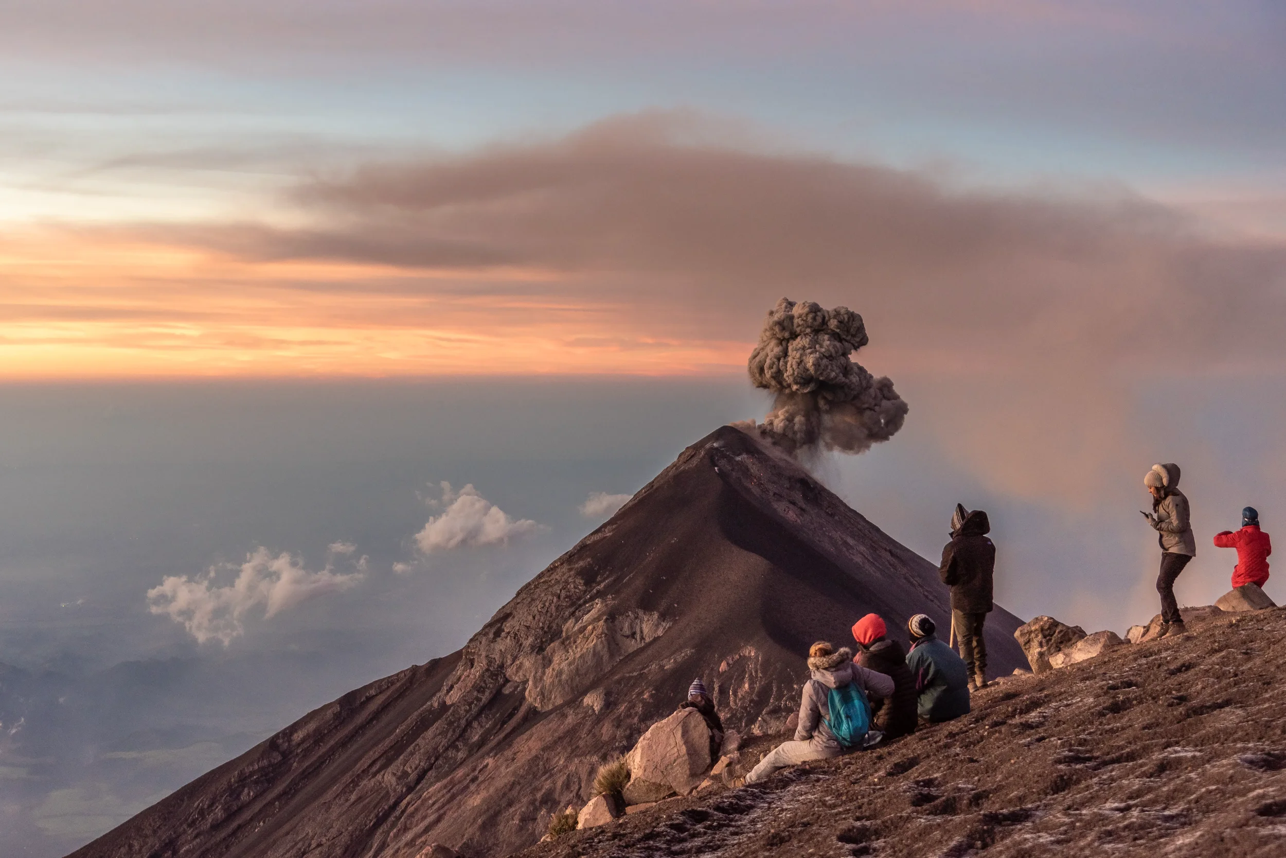 People standing and sitting on a rocky slope near a volcanic eruption, which emits ash and a large plume of smoke under a colorful sky at sunset or sunrise.