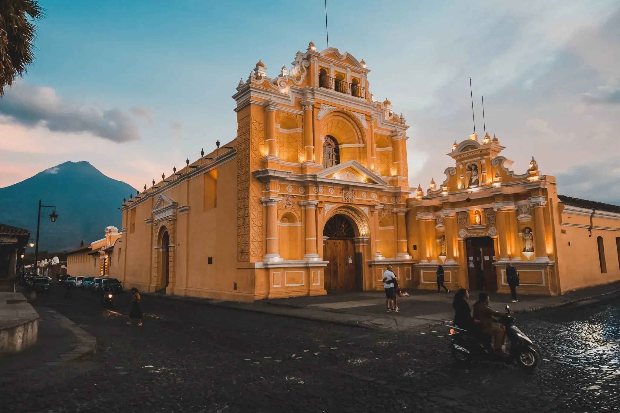 A yellow baroque-style church with intricate white trim details, located in a town plaza at dusk, with a mountain in the background.
