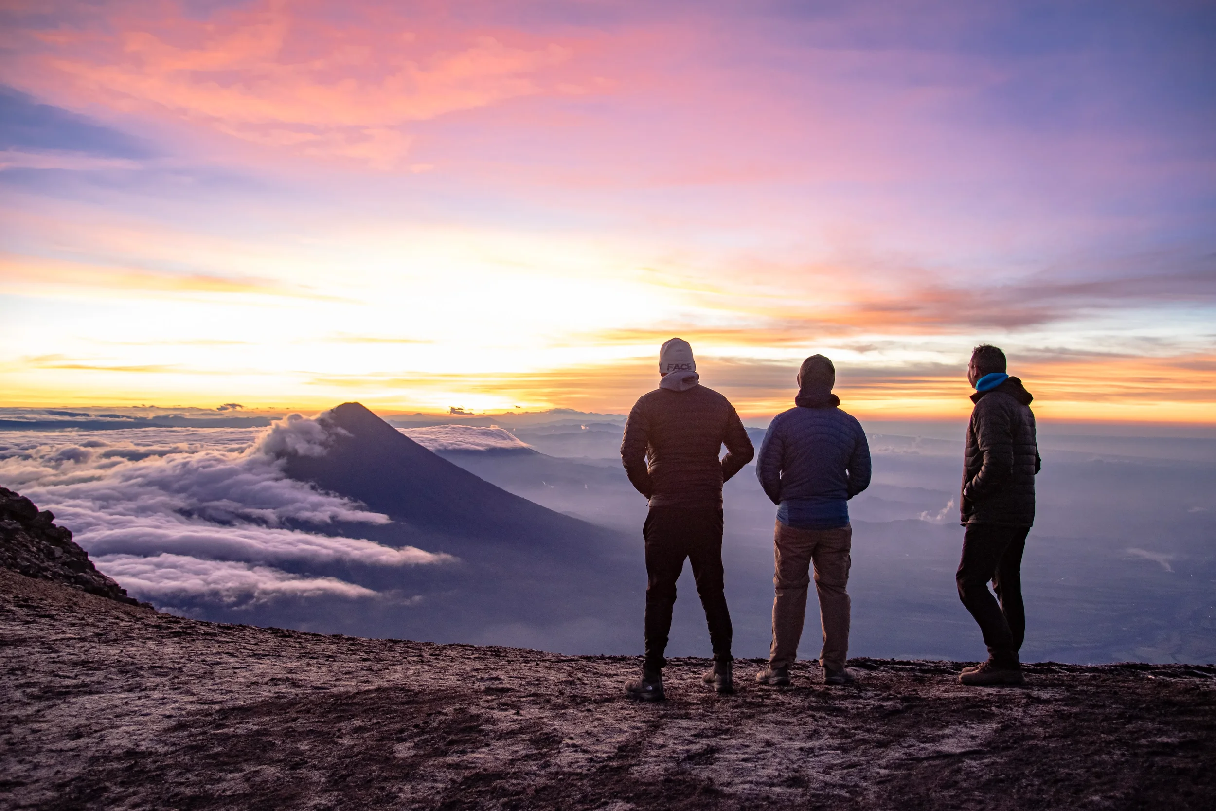 Three people are standing at Volcano Acatenango sunset, overlooking clouds and a distant volcano, with colorful sky and clouds creating a scenic view.