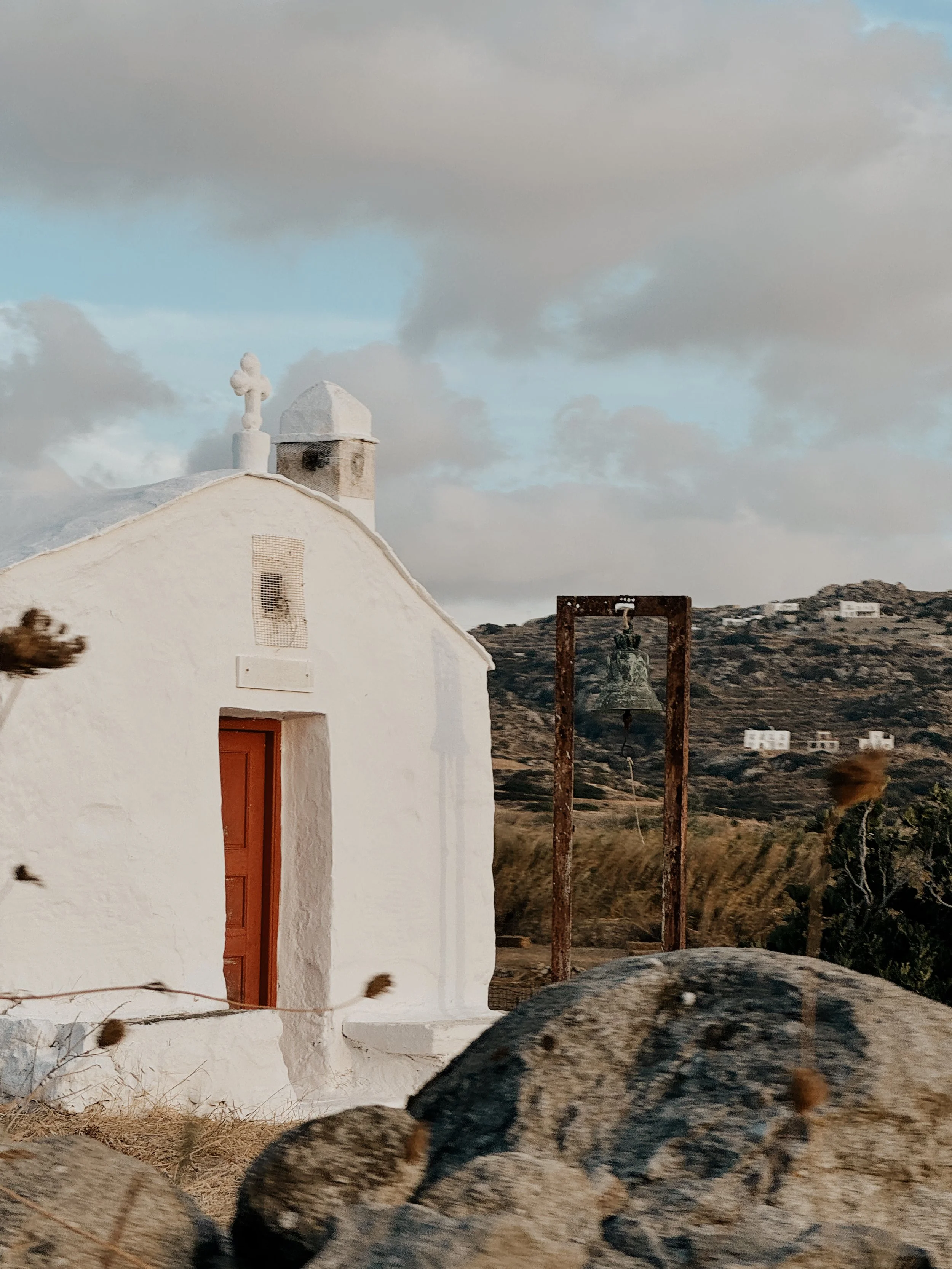 A white church with a bell tower and cross, set against a cloudy sky with hills and scattered houses in the background.