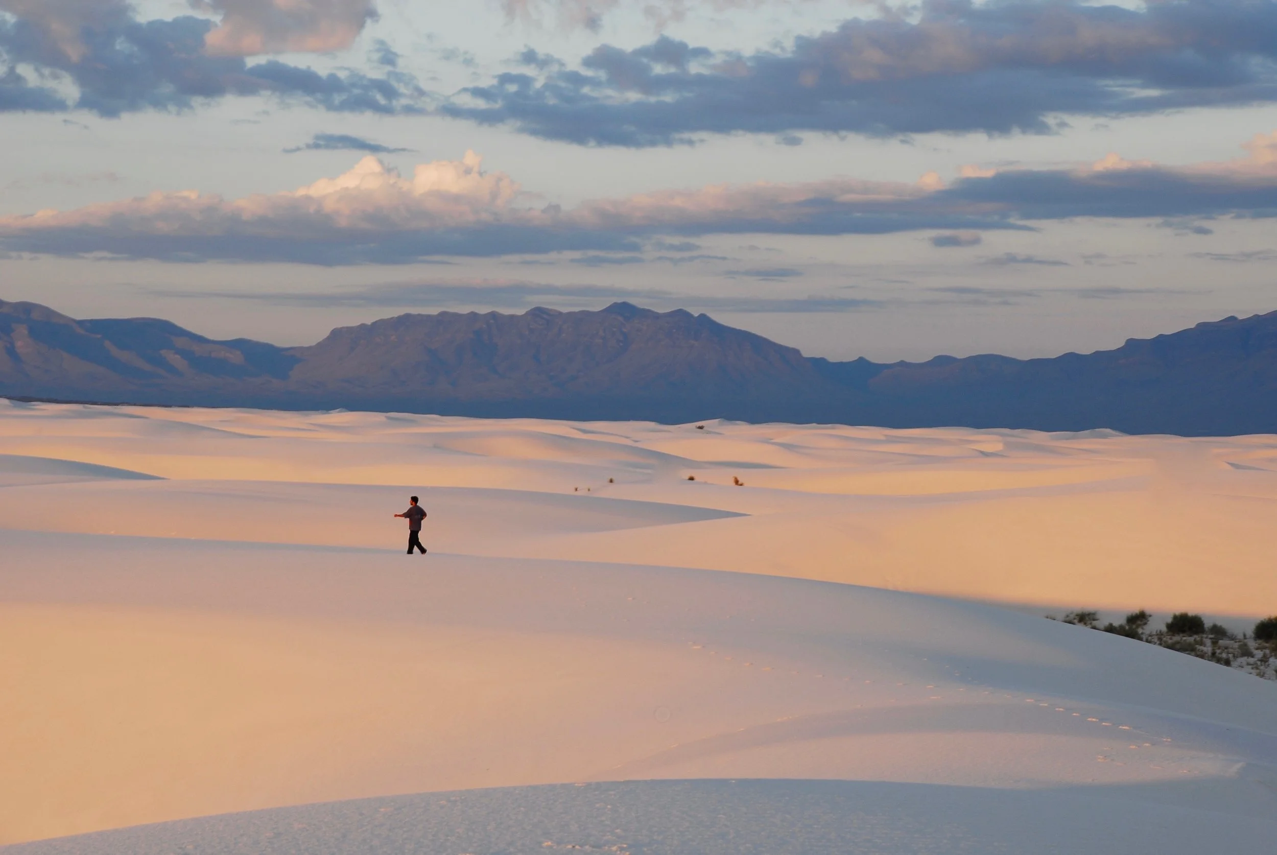 A man walking alone across a vast sandy desert with mountains in the background and a partly cloudy sky overhead.