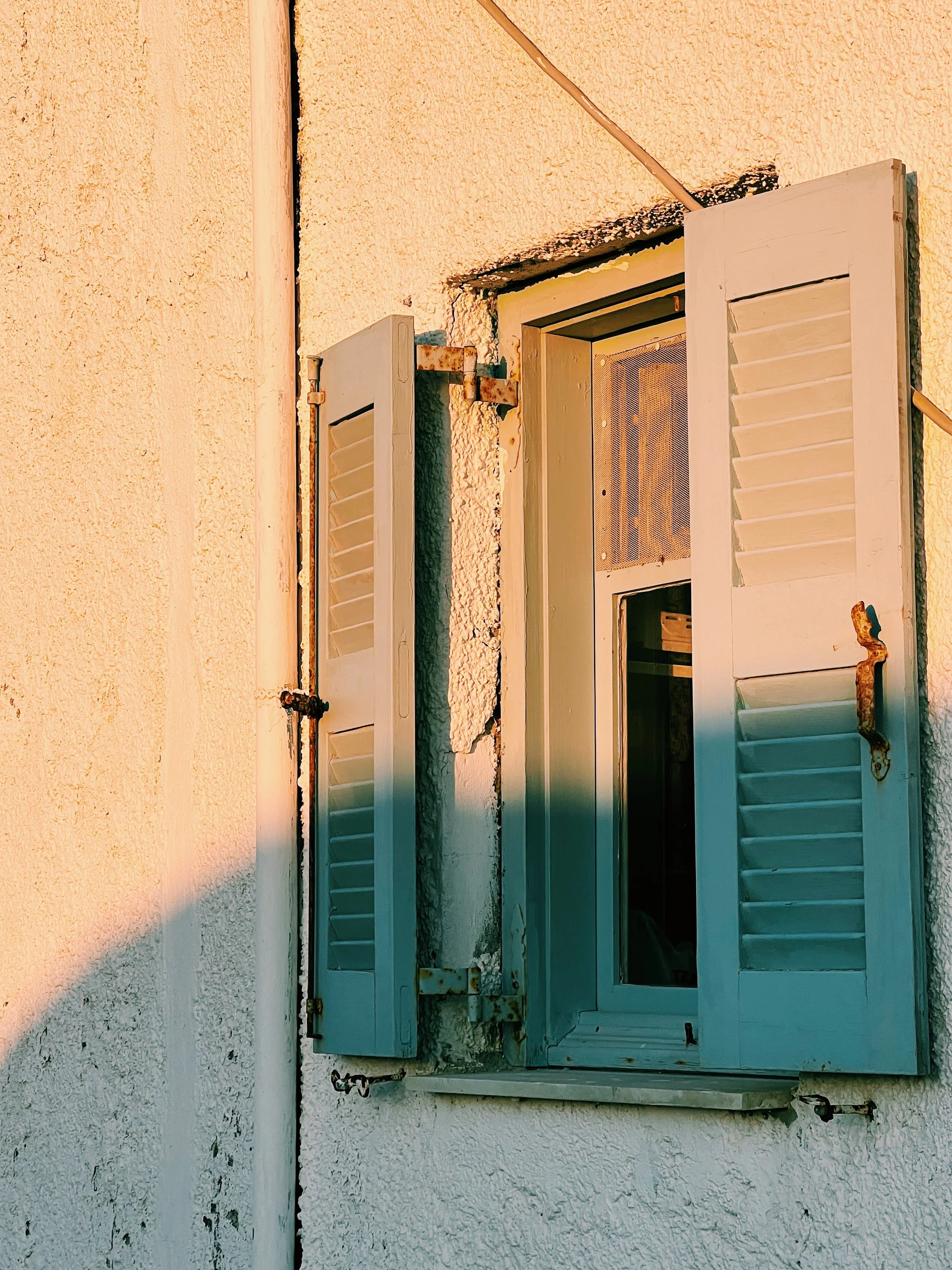 Close-up of an open window with white shutters on a white stucco wall, with rusted hinges and a rusty latch.