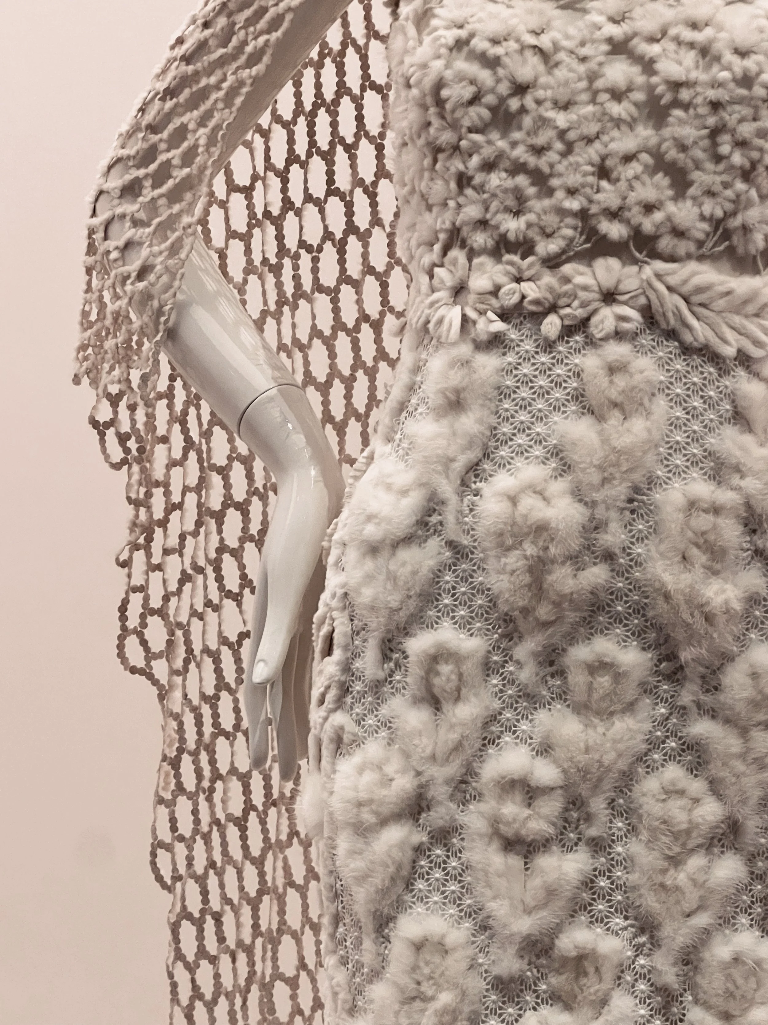 A close-up of a decorative white object with fuzzy and netted textures, including a beaded hanging element, against a plain background.
