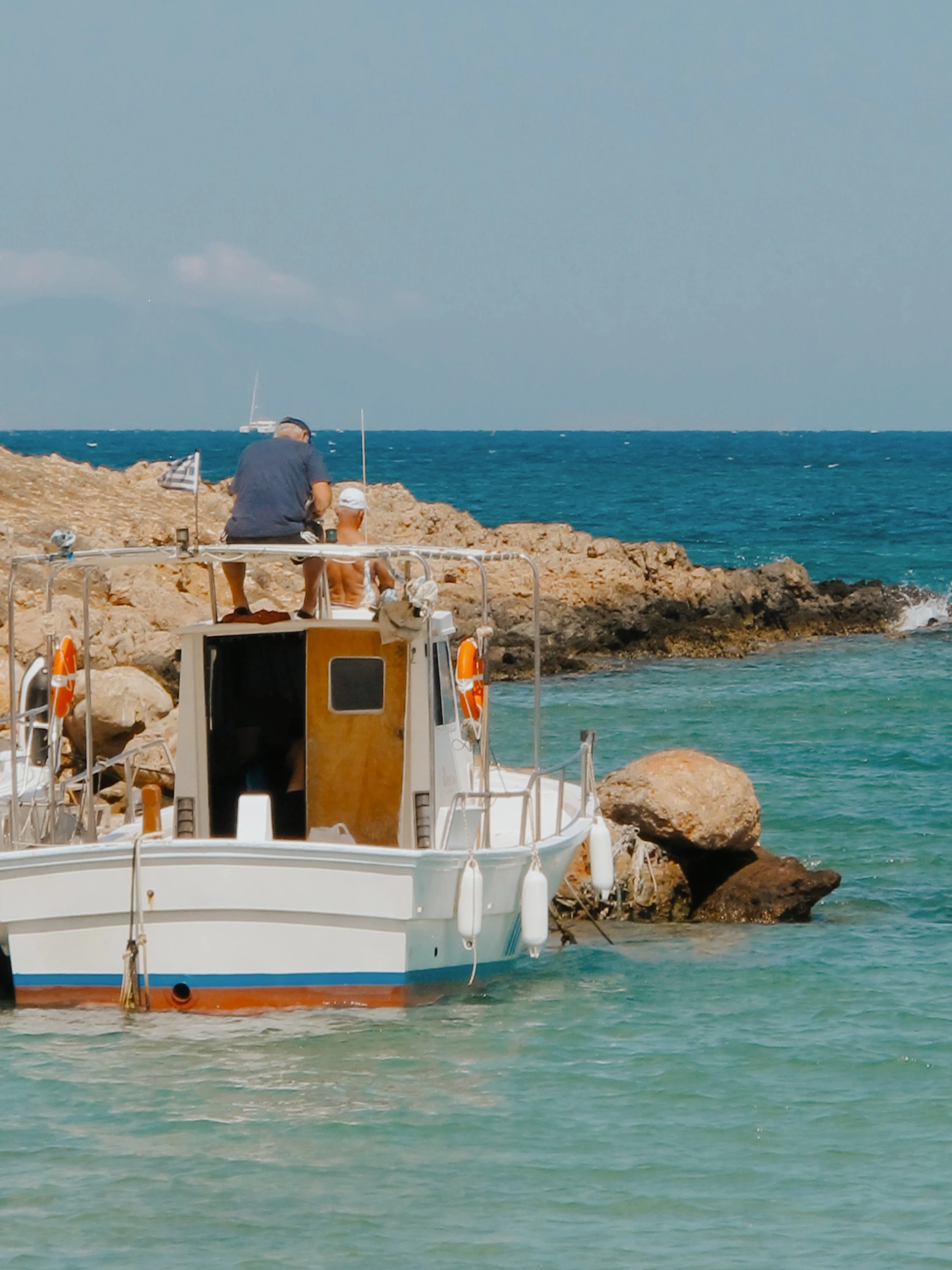 A boat docked near a rocky shoreline with two people on board, looking out over the ocean under a clear sky.