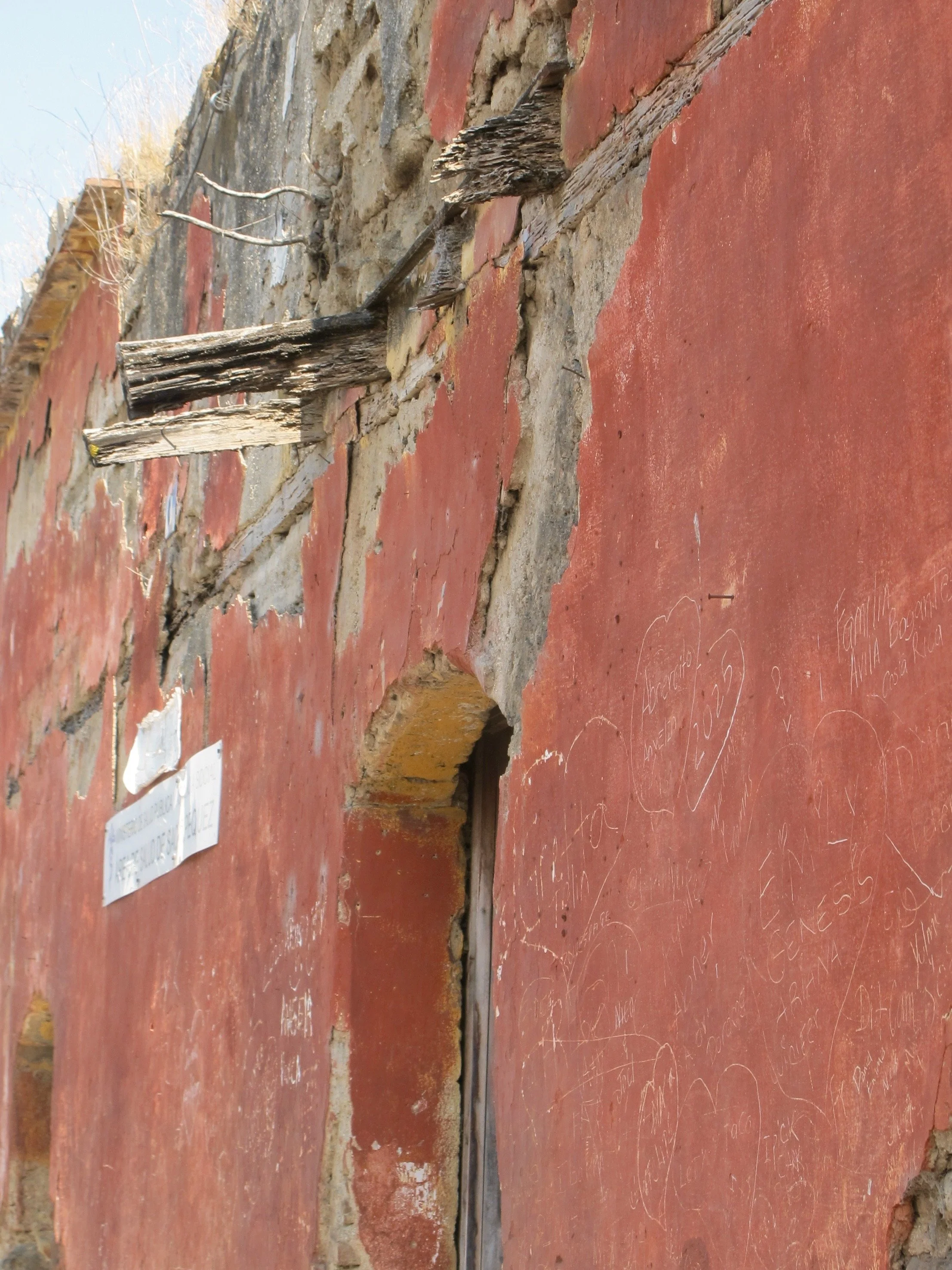 Close-up of a red brick wall with a narrow opening, possibly a window or doorway, with some handwritten notes and drawings on the wall. There are wooden and metal elements protruding from the wall, and a small sign with text is visible on the left si
