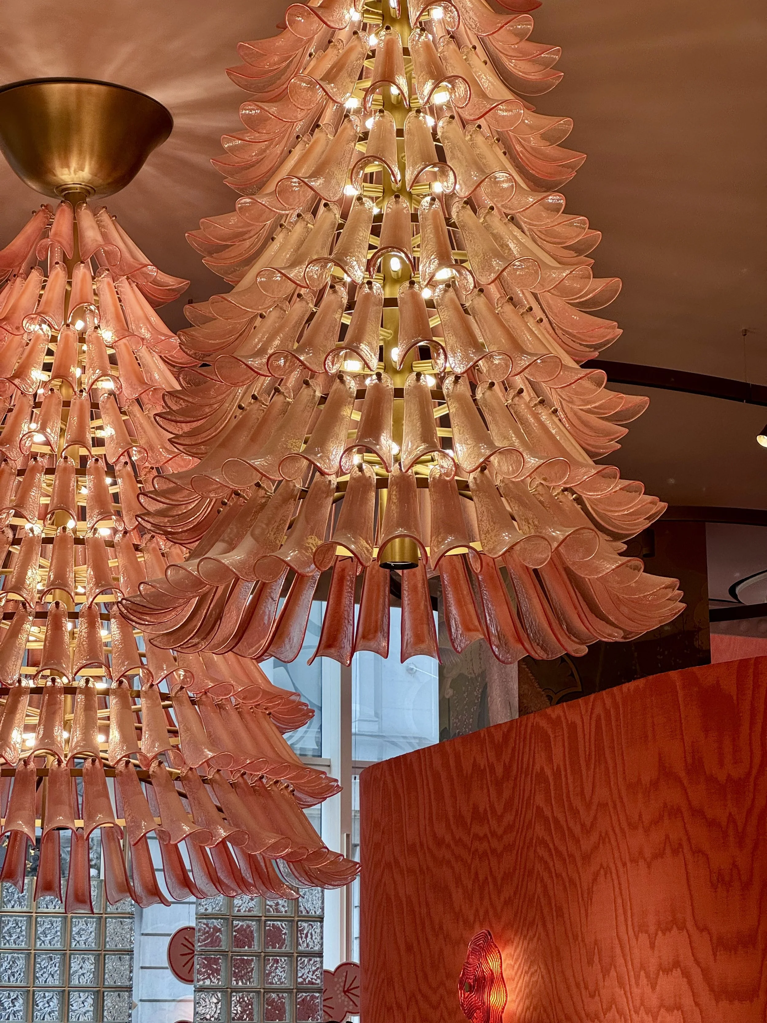 Close-up of a modern chandelier made of pink glass tubes with curved ends, arranged in a spherical shape, illuminated from within, hanging above a polished wooden surface in a room with glass block windows.