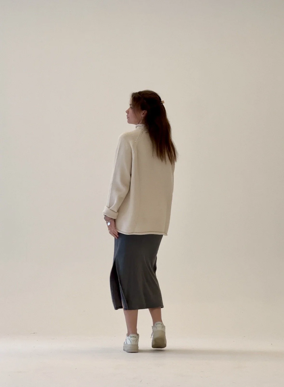 A woman standing in profile against a plain white background, wearing a cream-colored oversized jacket, dark midi skirt, and sneakers.