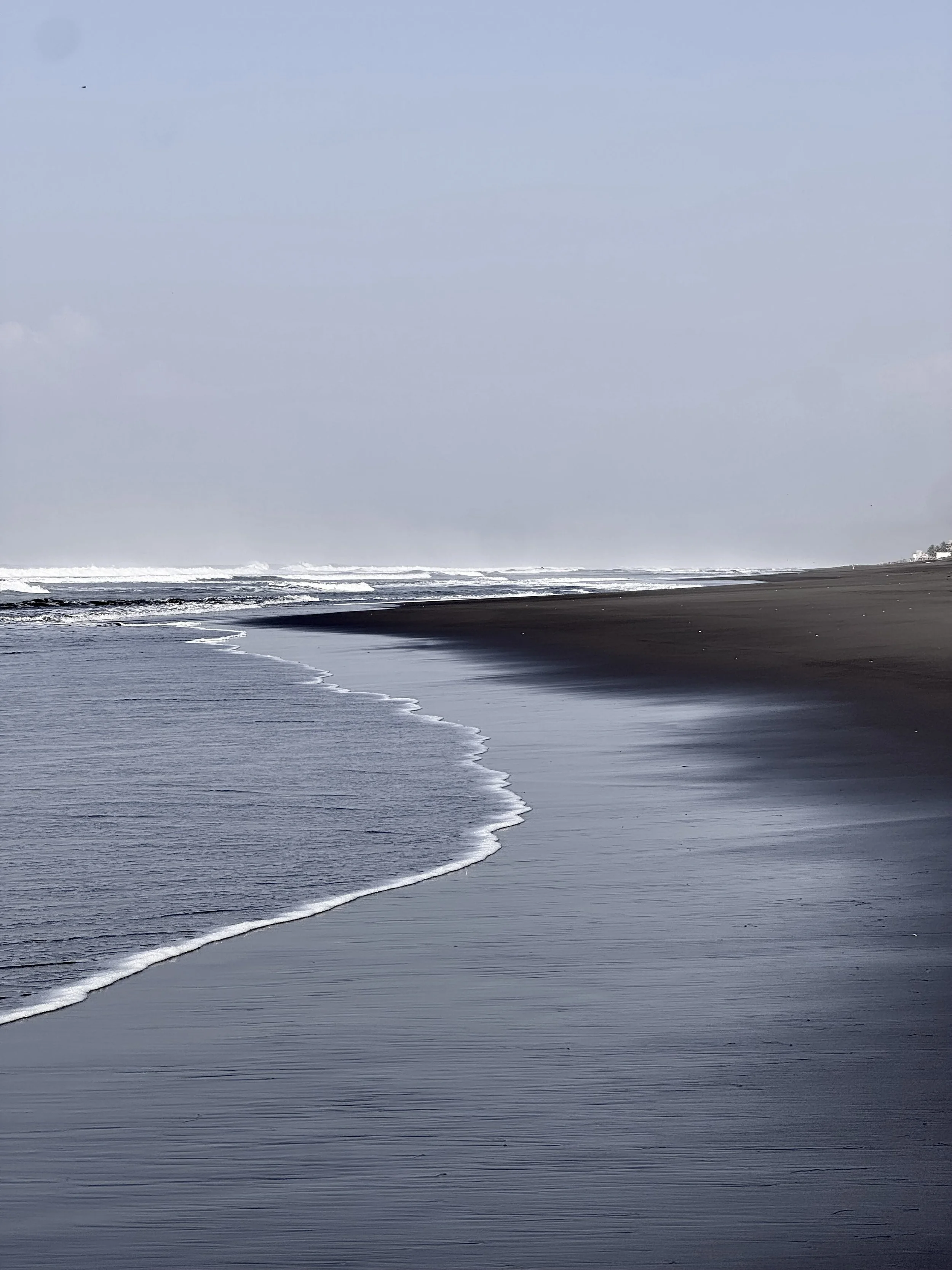 A dark sandy beach with gentle waves washing ashore under a cloudy sky.