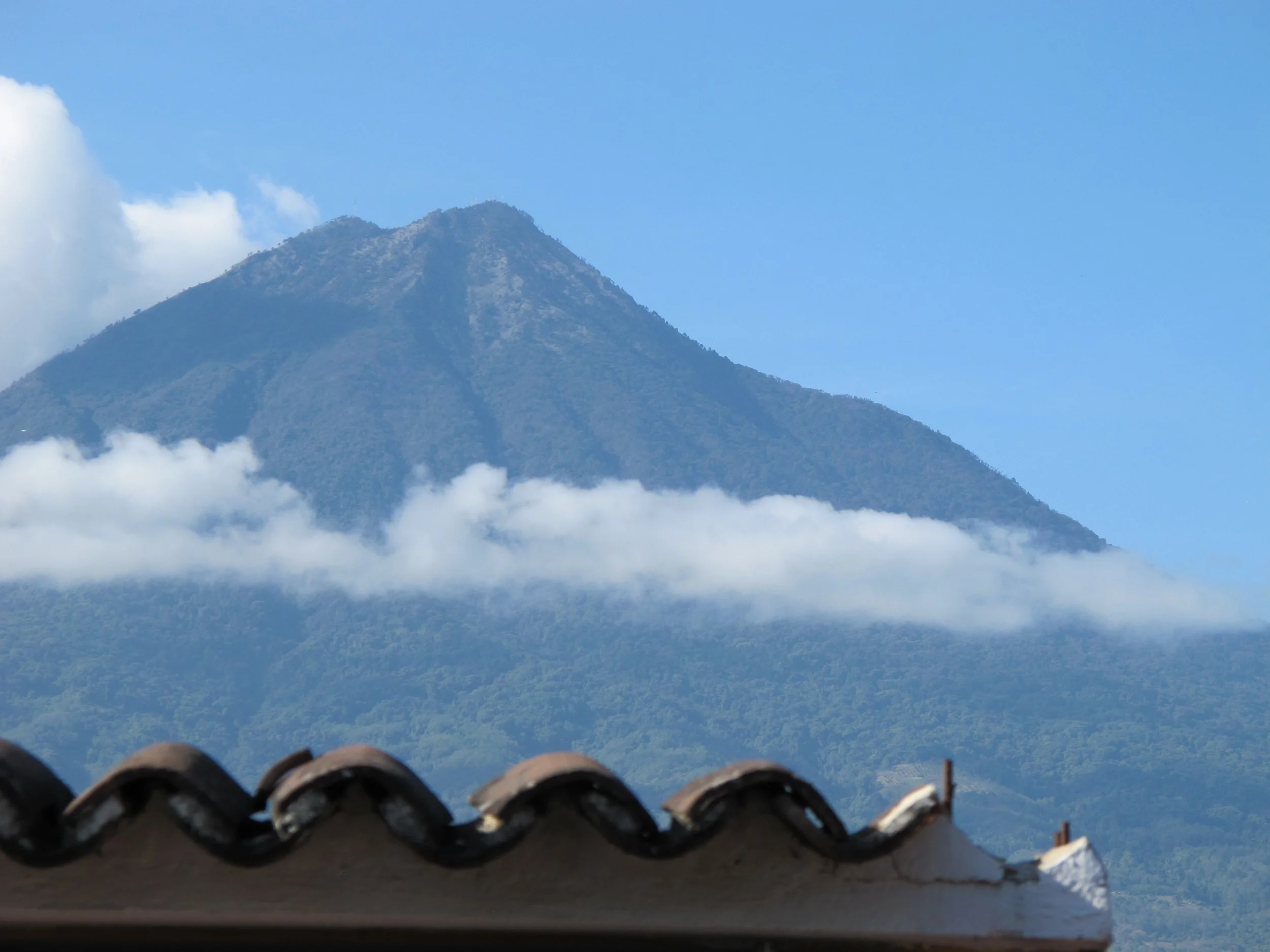 Mountaintop view with clouds at the base and a clear blue sky, viewed from a tiled roof.