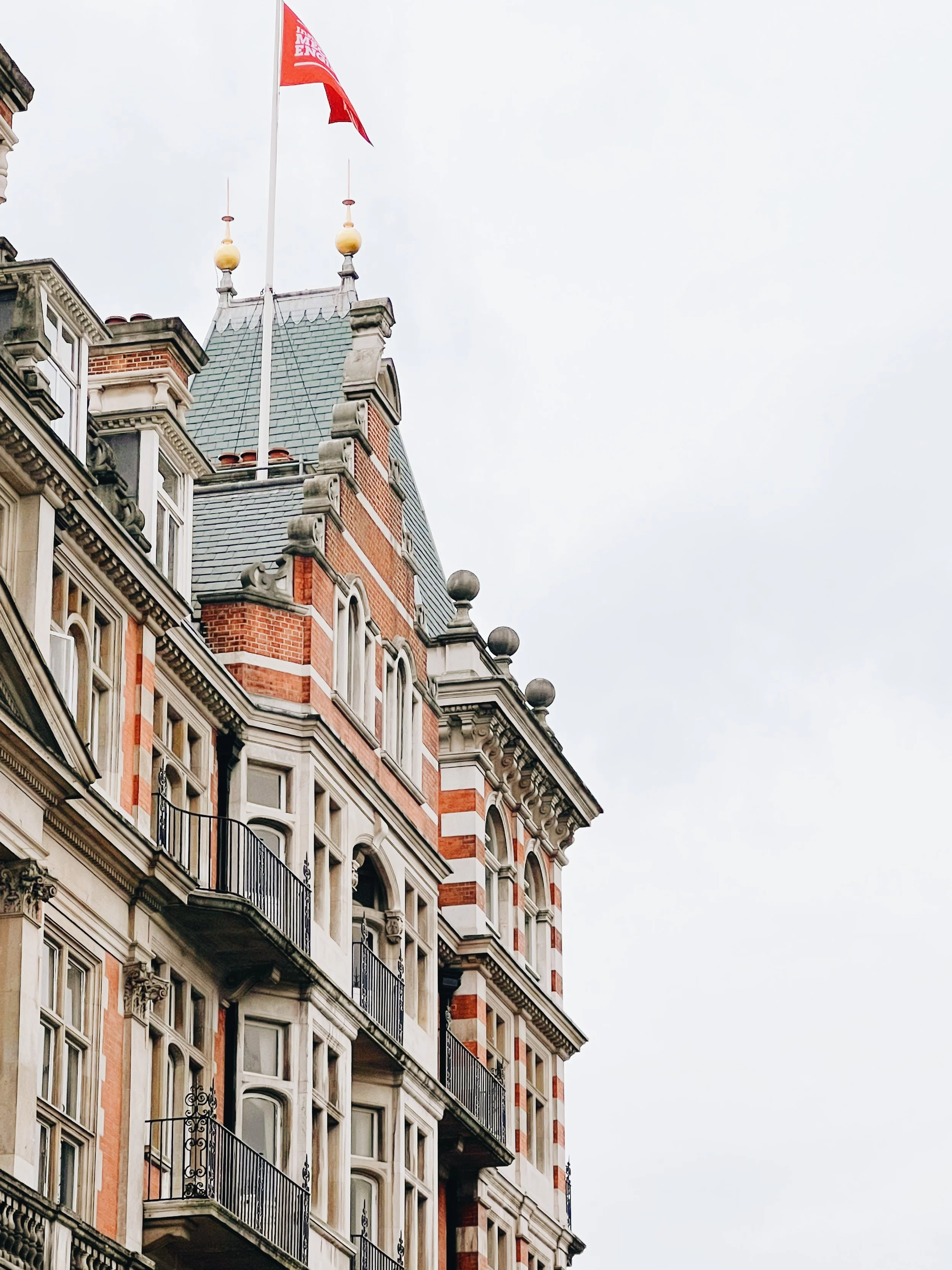 Historic building with ornate architecture, brick and stone facade, black wrought iron balconies, and a flagpole with a red flag on a cloudy sky background.