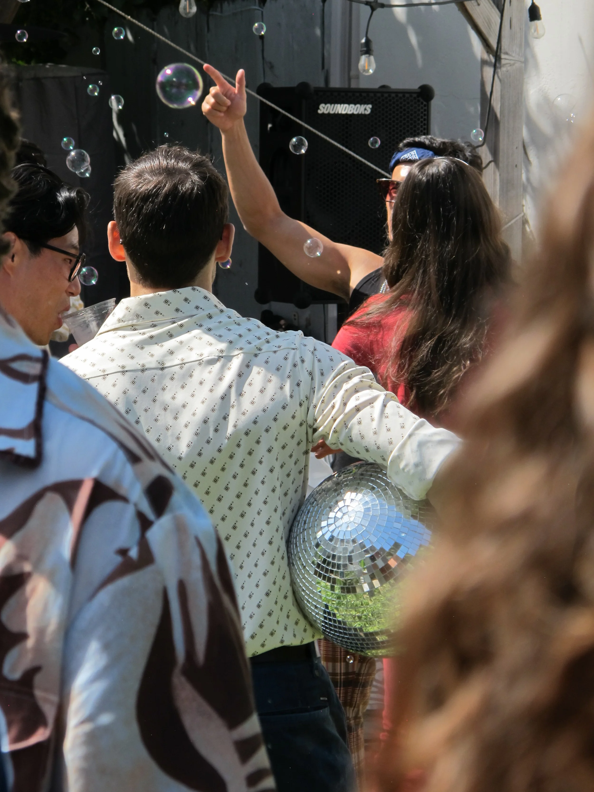 People at a party or gathering, with a woman in sunglasses and a blue bandana talking, surrounded by others, some holding drinks, and decorative string lights.