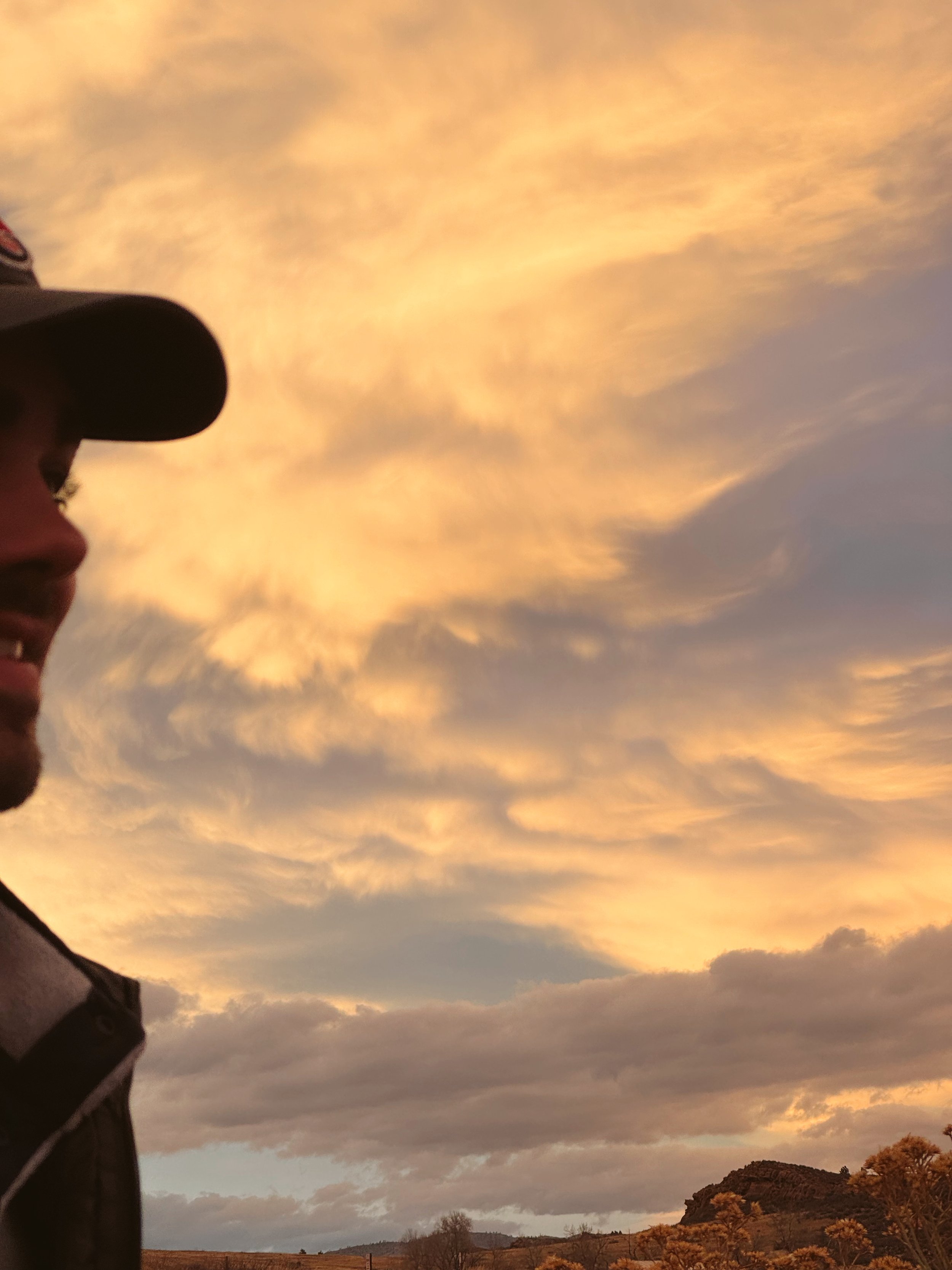 A person with glasses and a baseball cap looking at a sunset sky with clouds, overlooking a hilly landscape with trees.