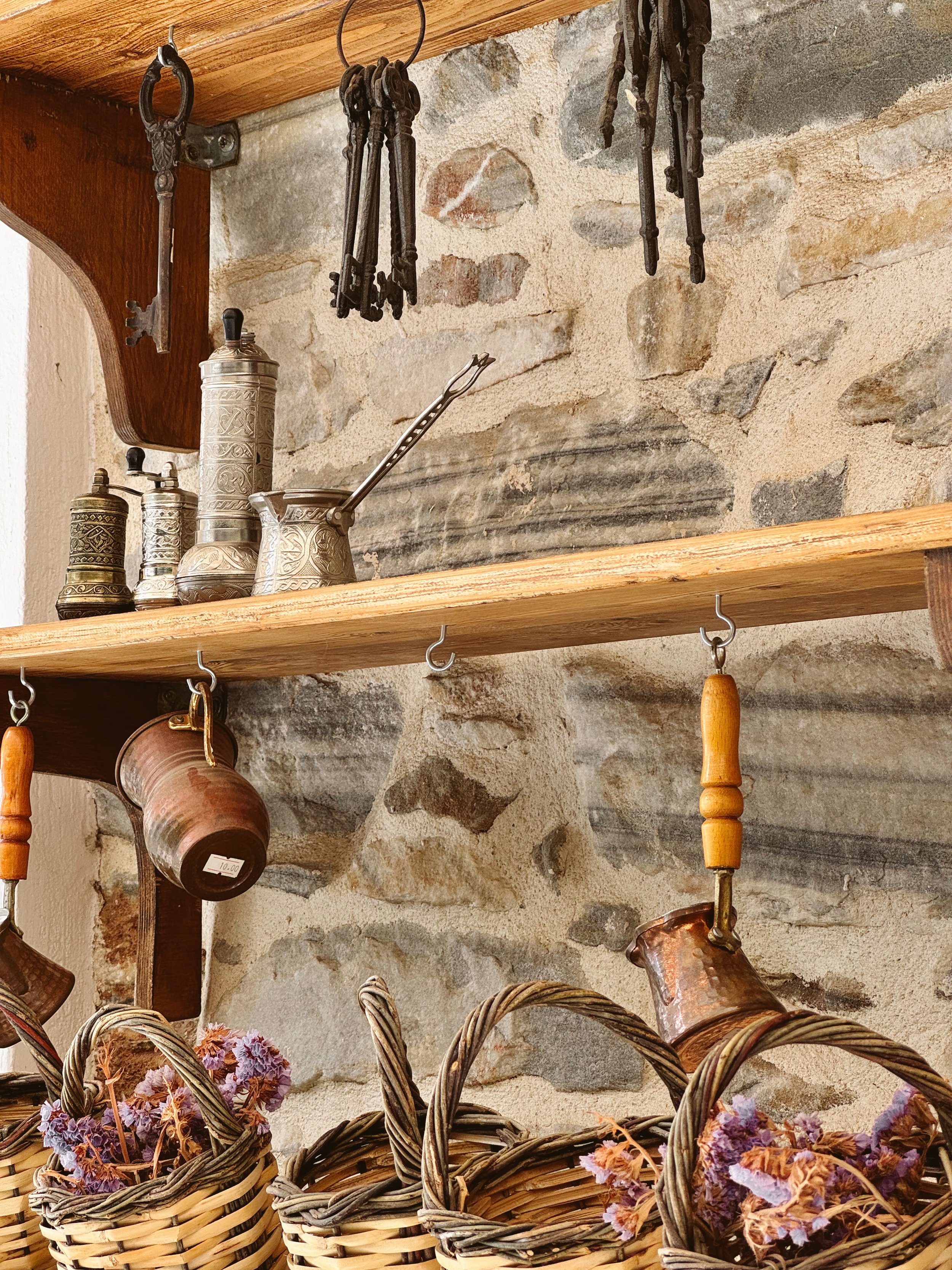 A rustic kitchen shelf holding vintage metal kitchen utensils and containers, with a stone wall in the background and woven baskets with dried flowers in the foreground.