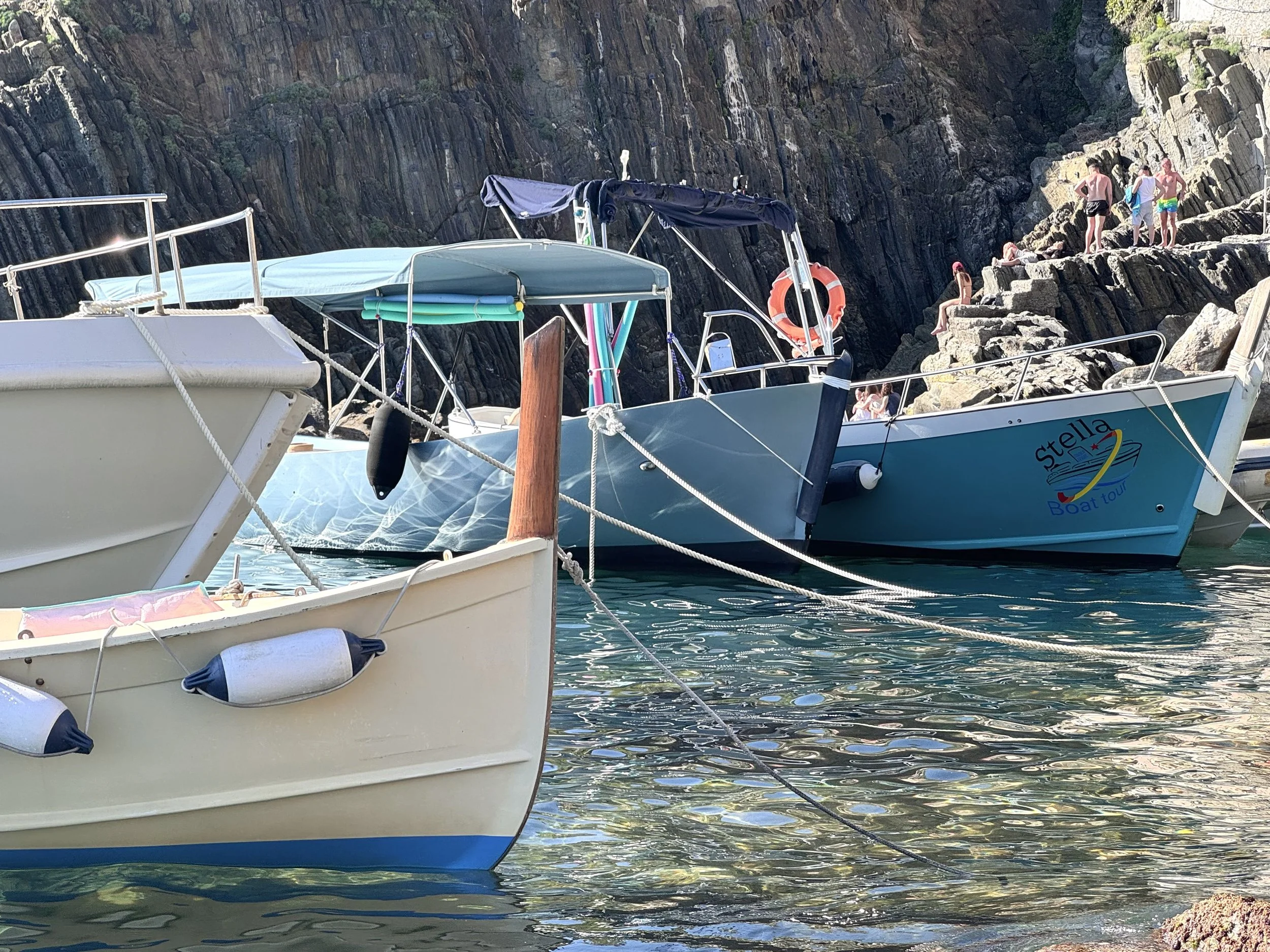 Boats docked in a rocky coastal area with people climbing rocks in the background.