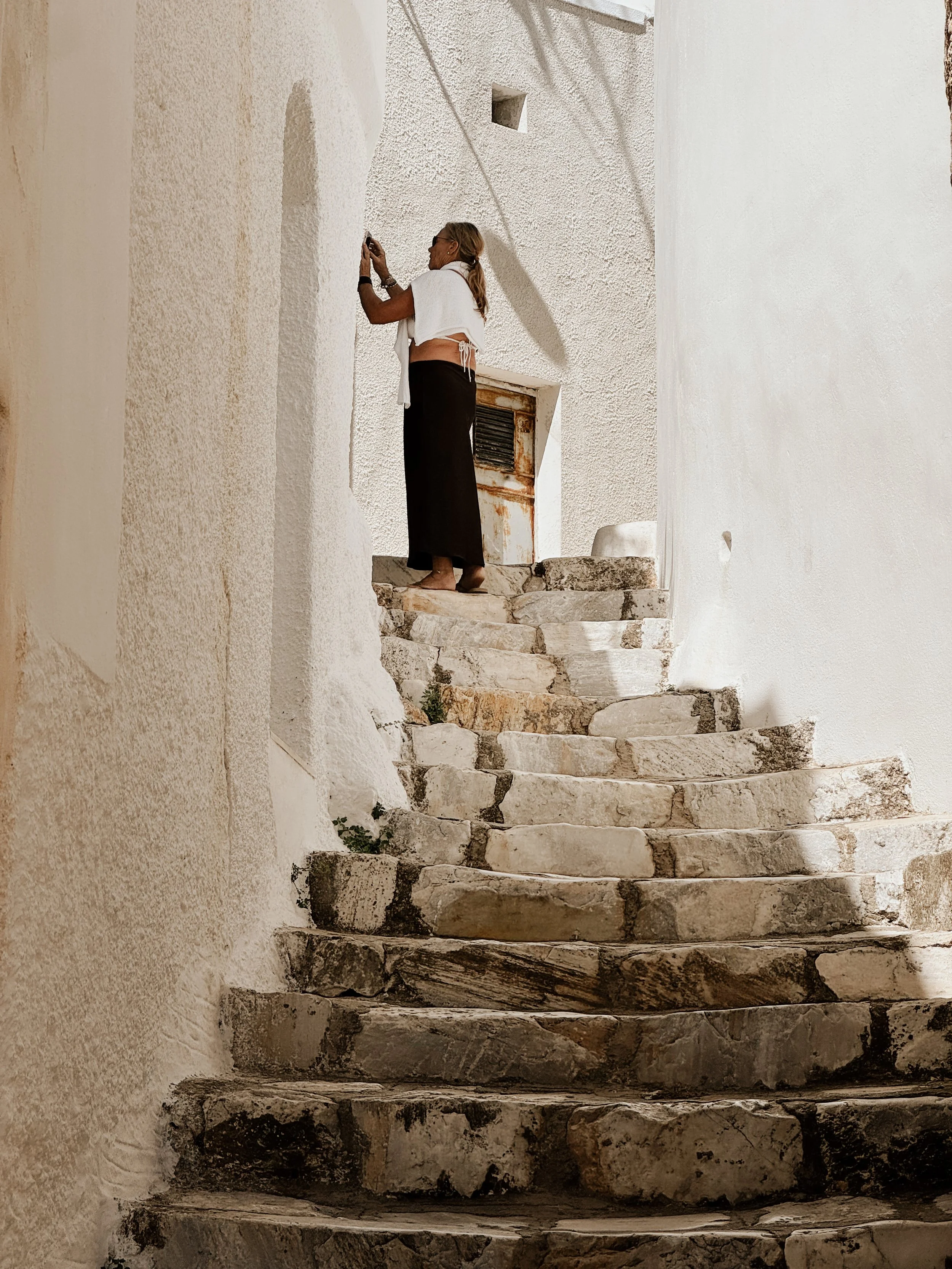 A woman with blonde hair, wearing glasses, a white top, and black pants, standing on stone stairs and taking a photo with her phone in a white-walled building with textured walls and rusted metal objects.