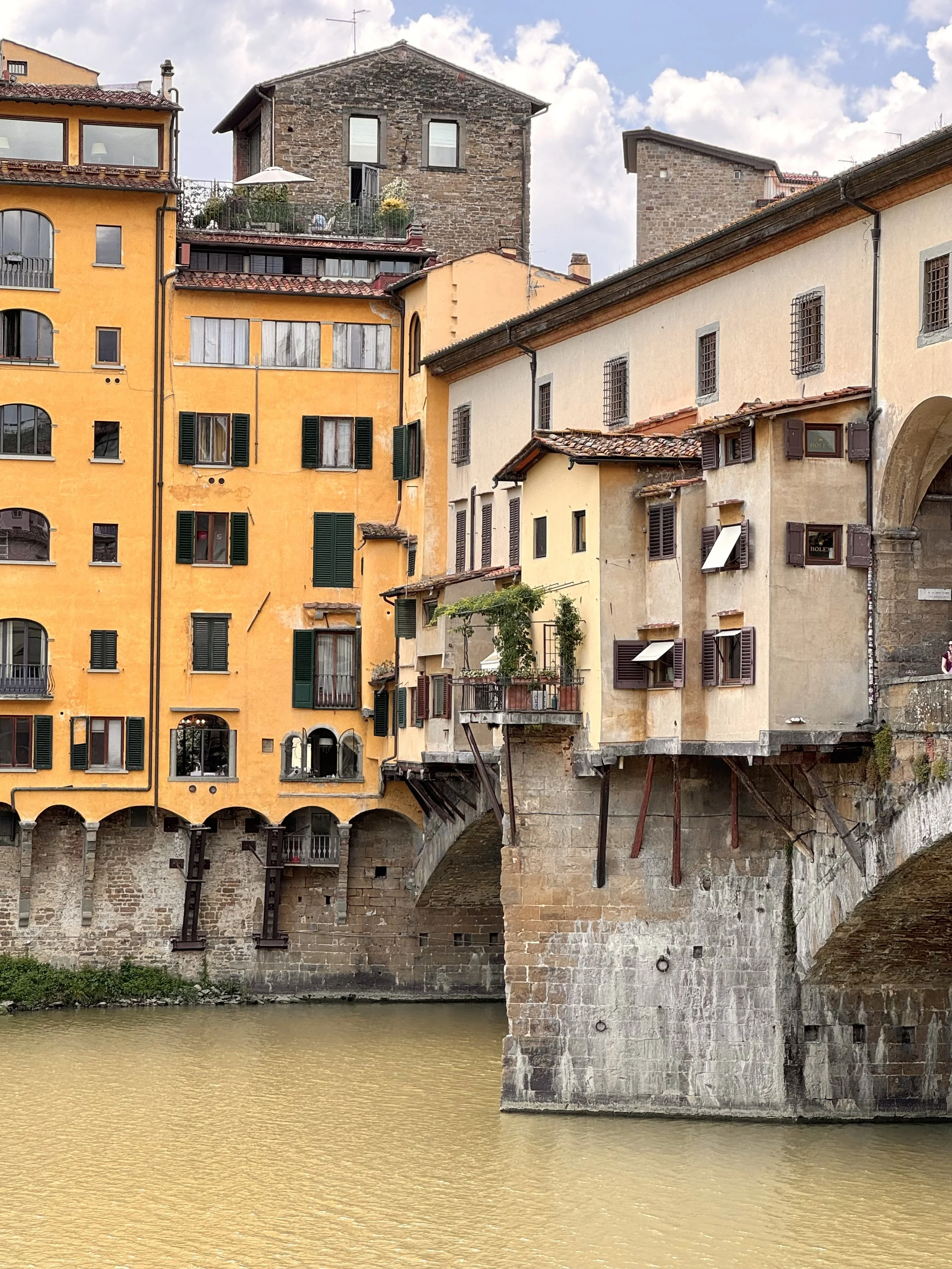 Colorful buildings with shuttered windows and balconies, built on a stone bridge over a river, with trees and sky in the background.