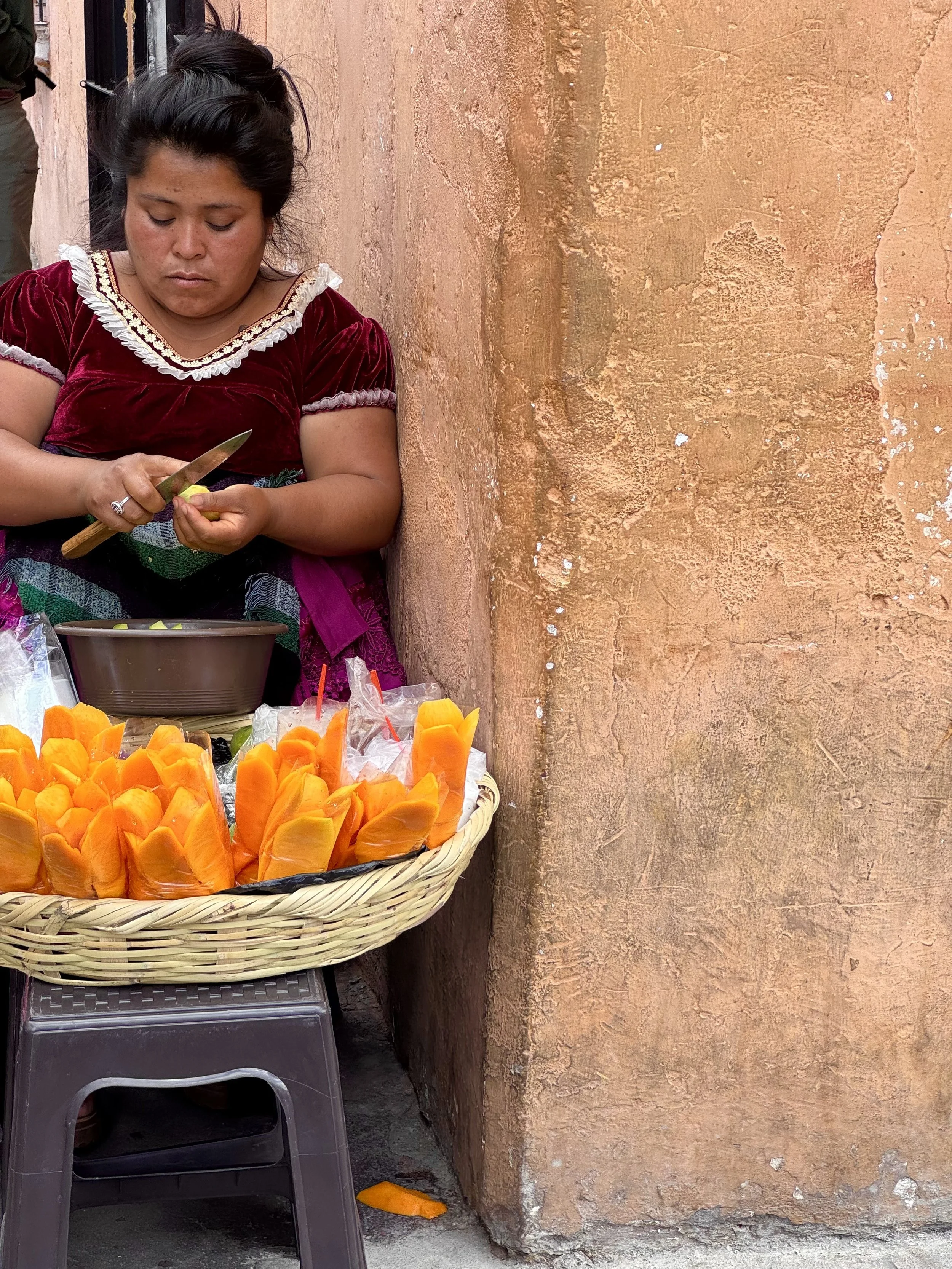A woman with dark hair tied up, wearing a red velvet dress with white frills on the neckline, sitting against an orange-brown textured wall, peeling fruit or preparing food, with a basket of orange and yellow papaya slices on a table in front of her.