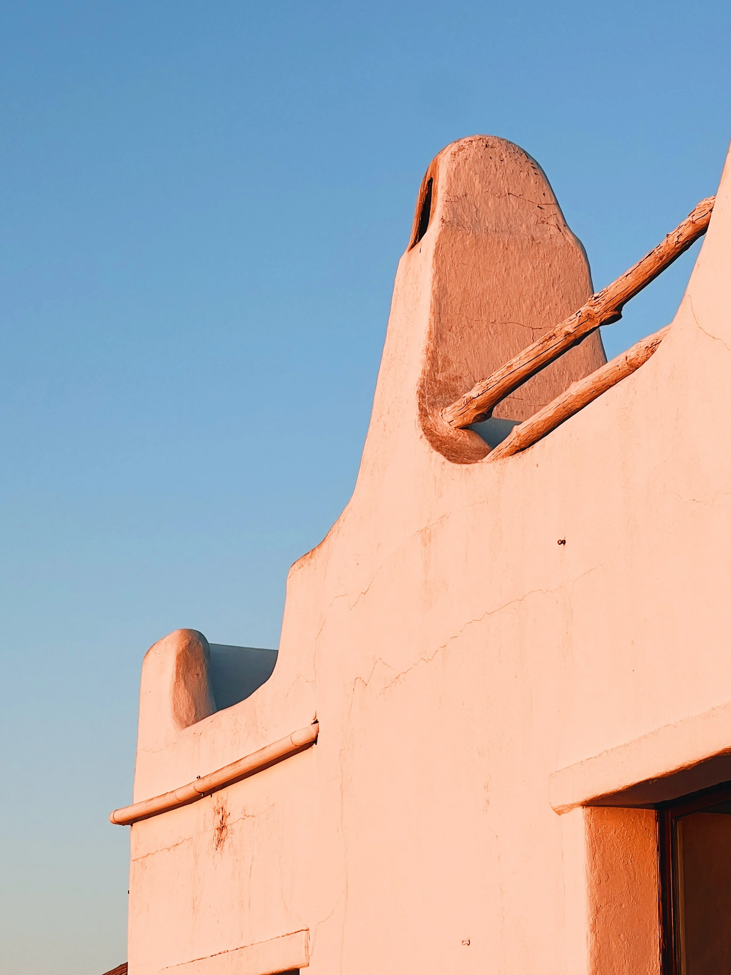 Close-up of a beige stucco building with wooden beams and a small window, against a clear blue sky.