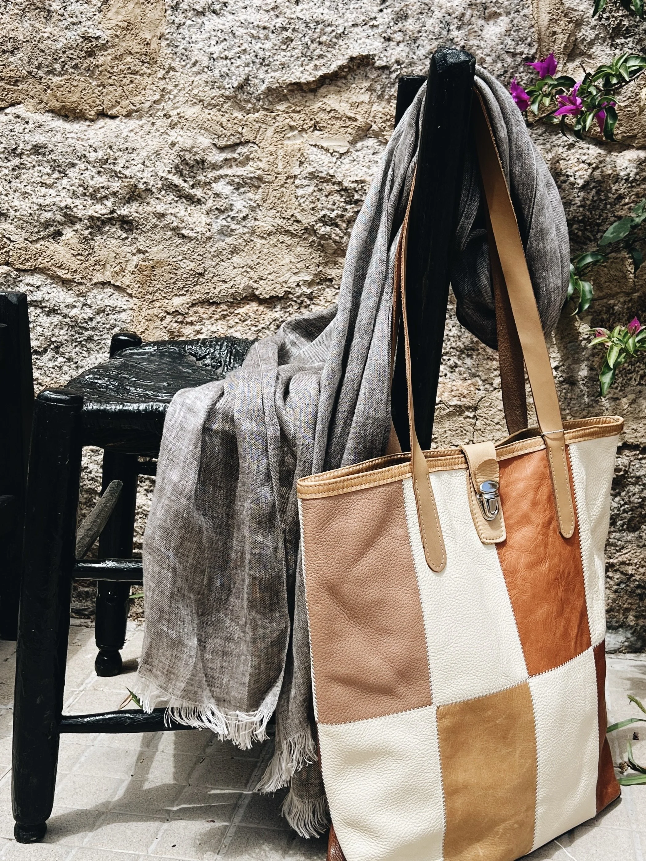 A black wooden chair with a scarf draped over the backrest, a large beige, white, and brown striped tote bag hanging from the side, a textured stone wall background, and purple flowers in the background.