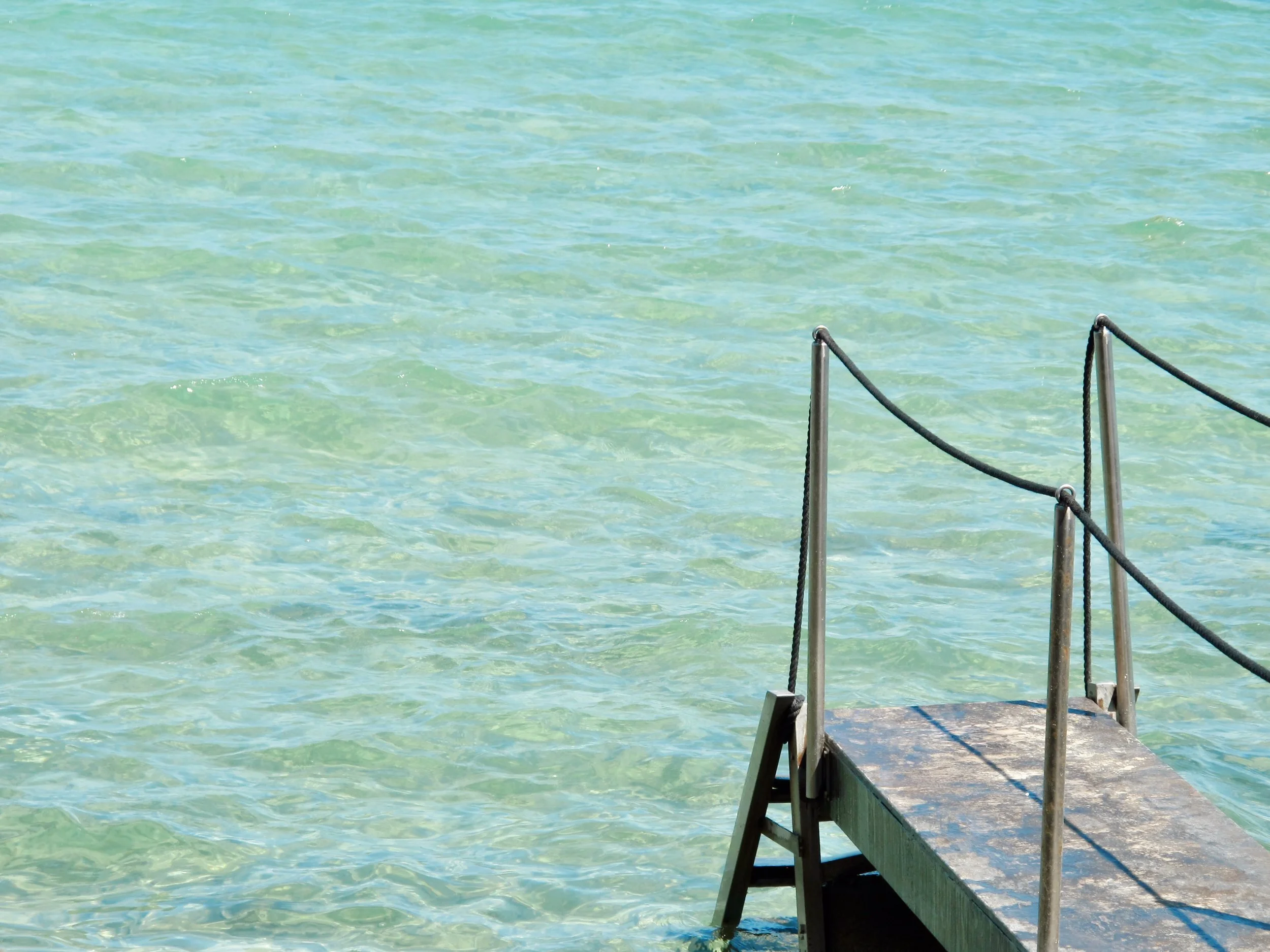 A wooden dock with metal railing extends into the clear, greenish water of a lake or ocean, with gentle waves.