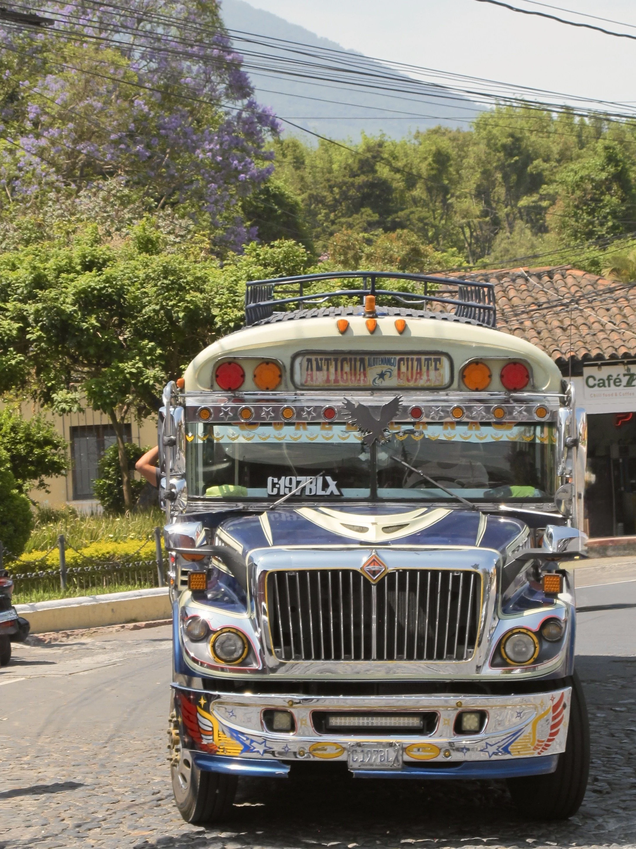 A decorated truck with colorful designs, chrome accents, and a license plate from Guatemala parked on a cobblestone street with trees and buildings in the background.