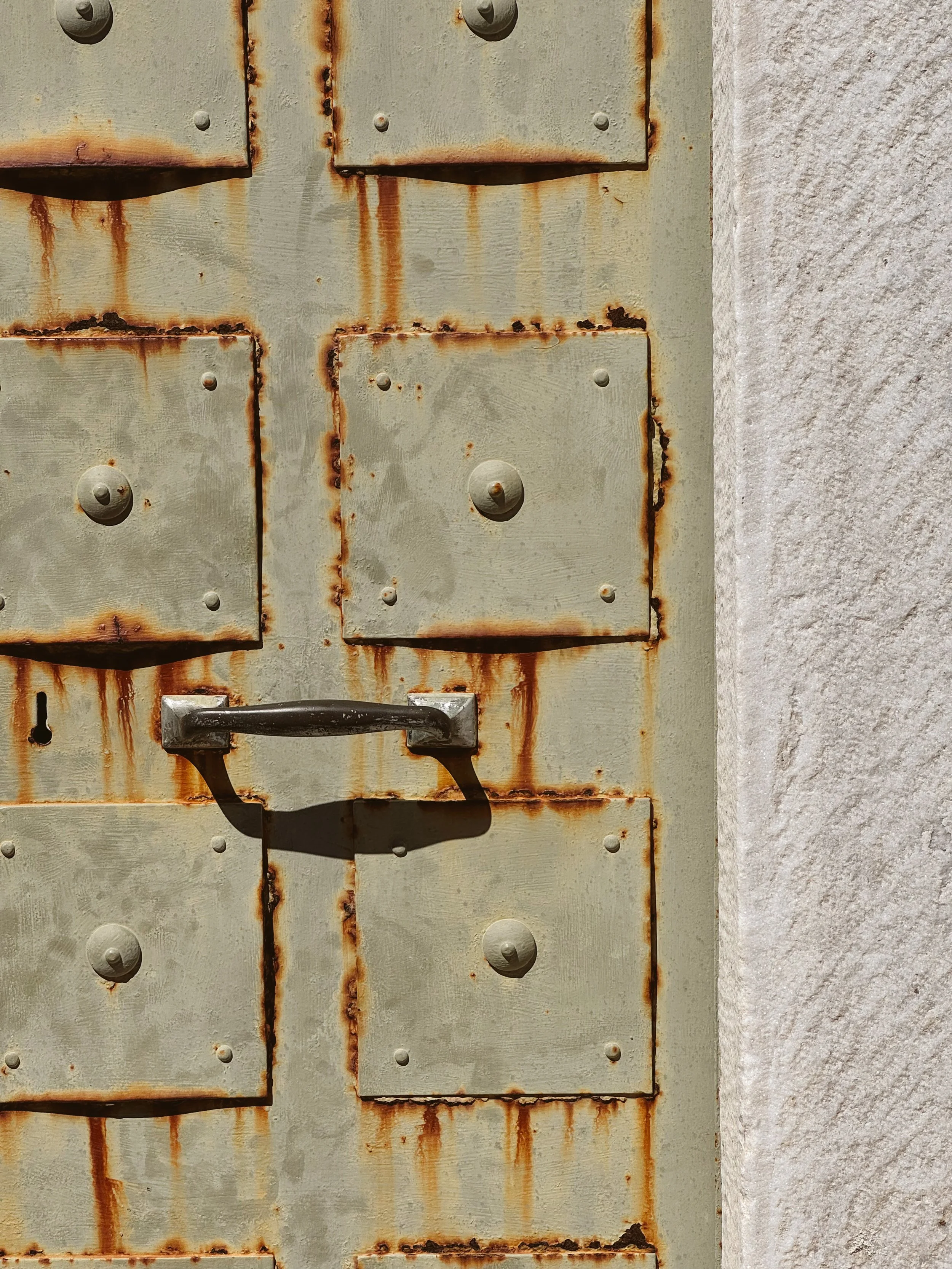 Close-up of a rusty, weathered metal door with a black handle and visible bolt heads, joined to a rough concrete wall.
