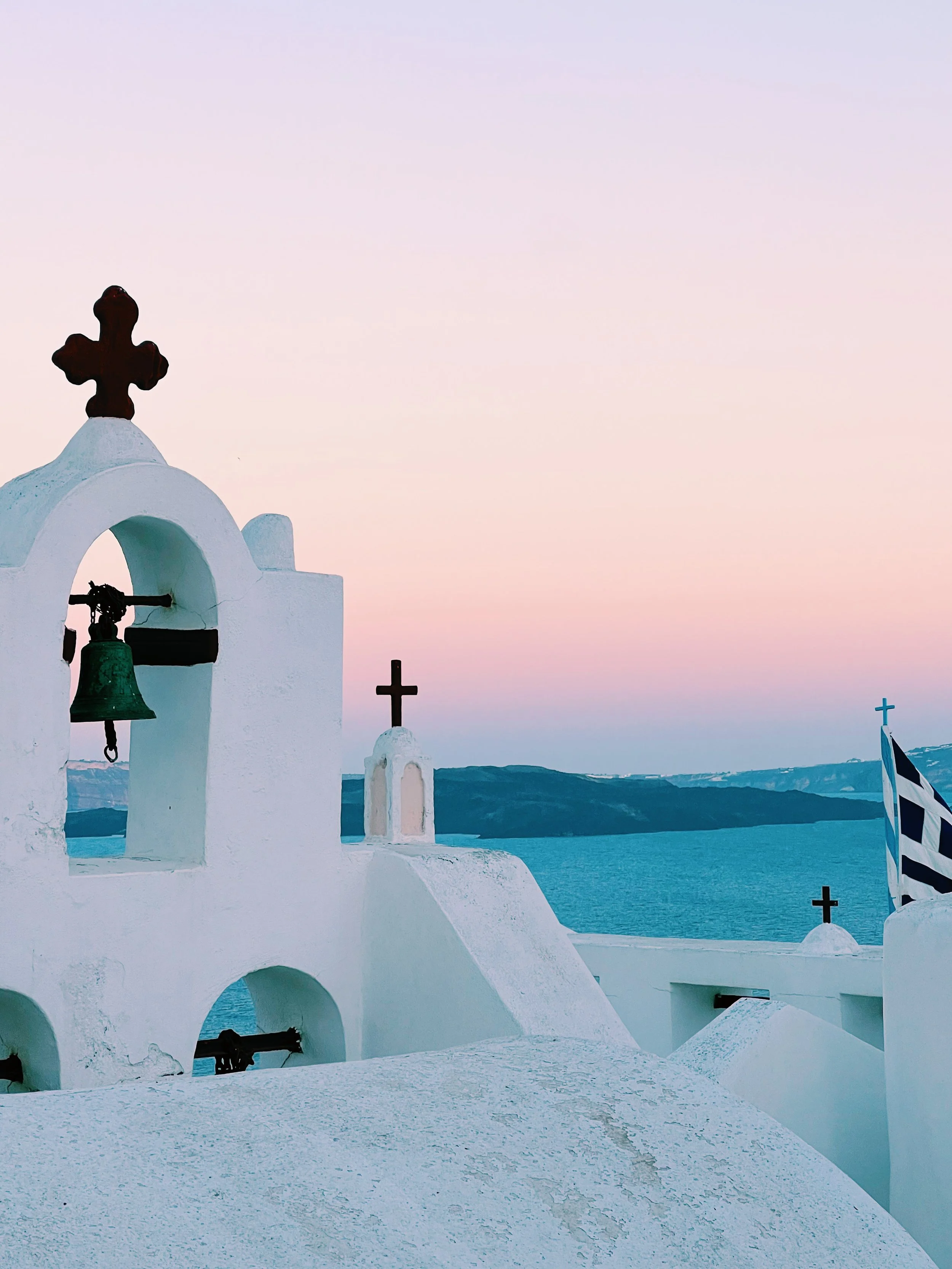White buildings with arches and crosses, blue sky and water in the background, typical of Greek island architecture, at sunset.