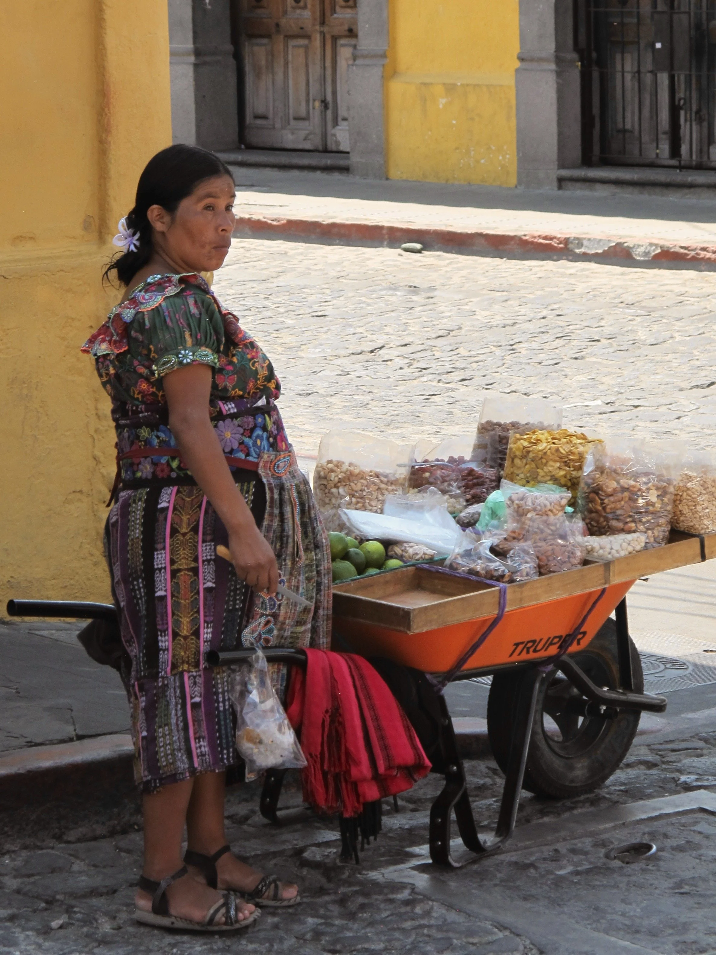 A woman wearing traditional clothing and sandals stands beside a small cart filled with bags of snacks and candies on a cobblestone street, with a yellow wall and wooden door in the background.