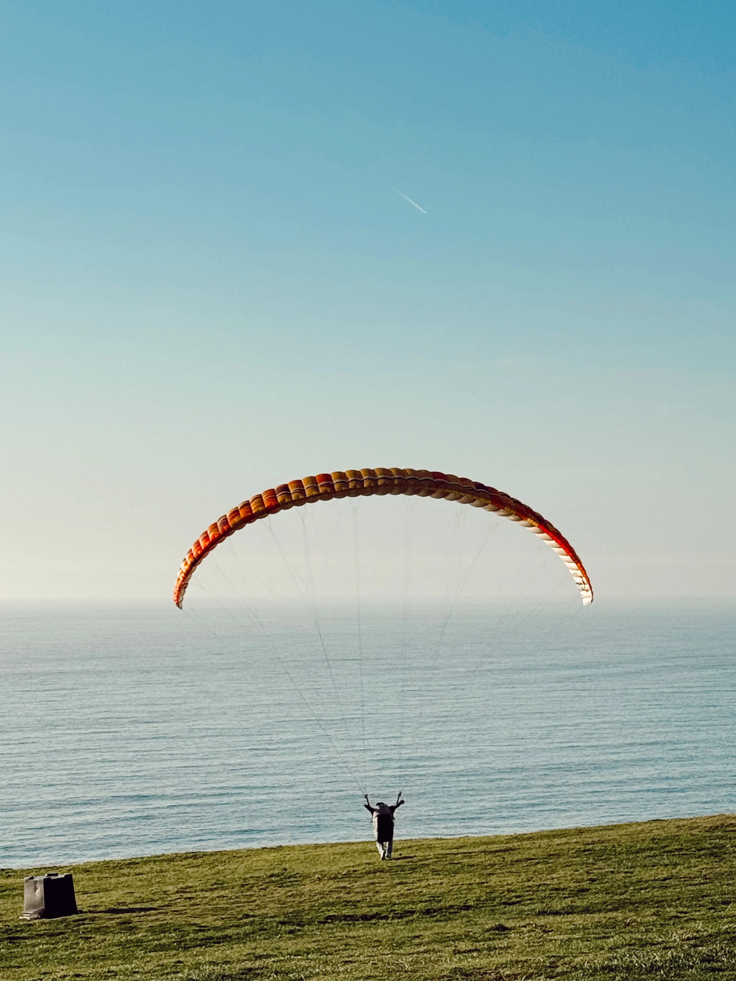 Person flying a paramotor along a coastline with grass and water, sky in background.