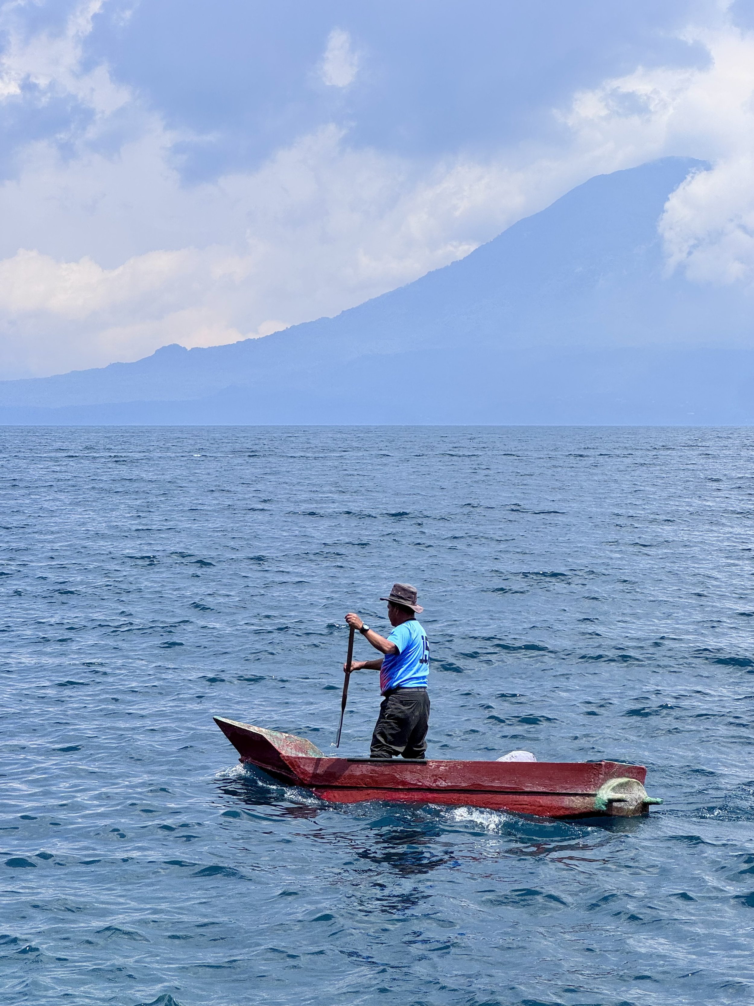 Man standing on a small red boat, wearing a hat and casual clothing, paddling on a calm body of water with mountain and cloudy sky in the background.